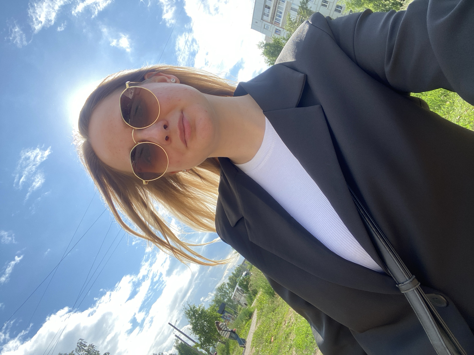 A young woman with blonde hair and sunglasses is taking a selfie outdoors on a sunny day. She is wearing a black blazer over a white top and a black crossbody bag. In the background, there is a green grassy area, trees, a sidewalk, and a multi-story building. The sky is blue with white clouds and the sun is shining brightly. A person in a striped shirt and jeans is walking on the sidewalk in the distance. The location appears to be an urban environment in Chaikovsky, Russia, given the presence of apartment buildings and utility poles.
