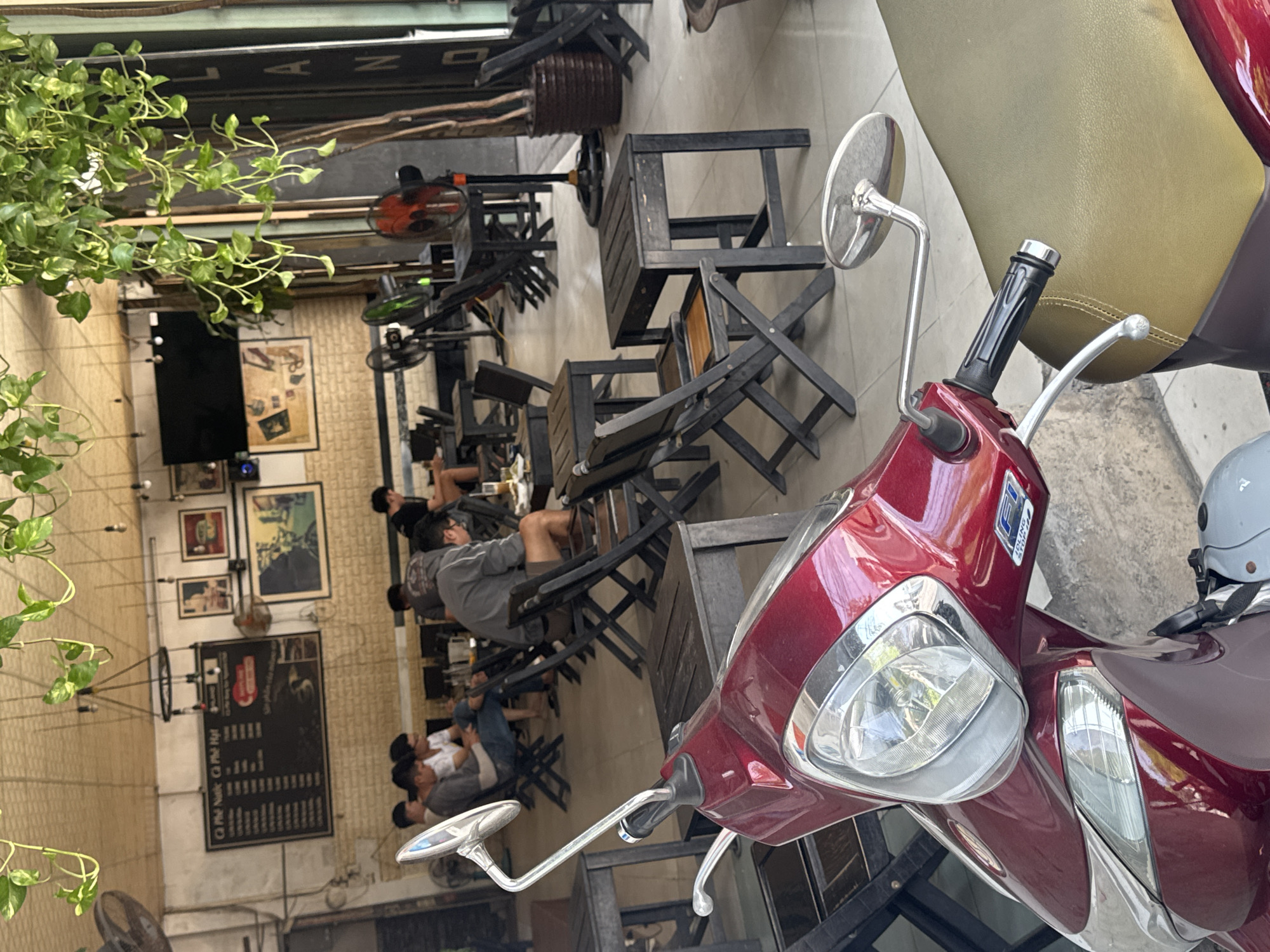 This is an eye-level, medium shot of a coffee shop in Nha Trang, Vietnam. In the foreground, a red scooter is parked with its front end angled towards the viewer. The scooter has a shiny chrome mirror extending from its handle. The background shows the interior of a cafe with several patrons seated at dark wooden tables and chairs. Some individuals are engaged in conversations or looking at their phones, while others are seen from behind. The cafe is decorated with hanging plants and framed pictures on the wall. Various fans are positioned around the room. A large blackboard menu is visible on the wall behind the patrons. The lighting suggests it is daytime.