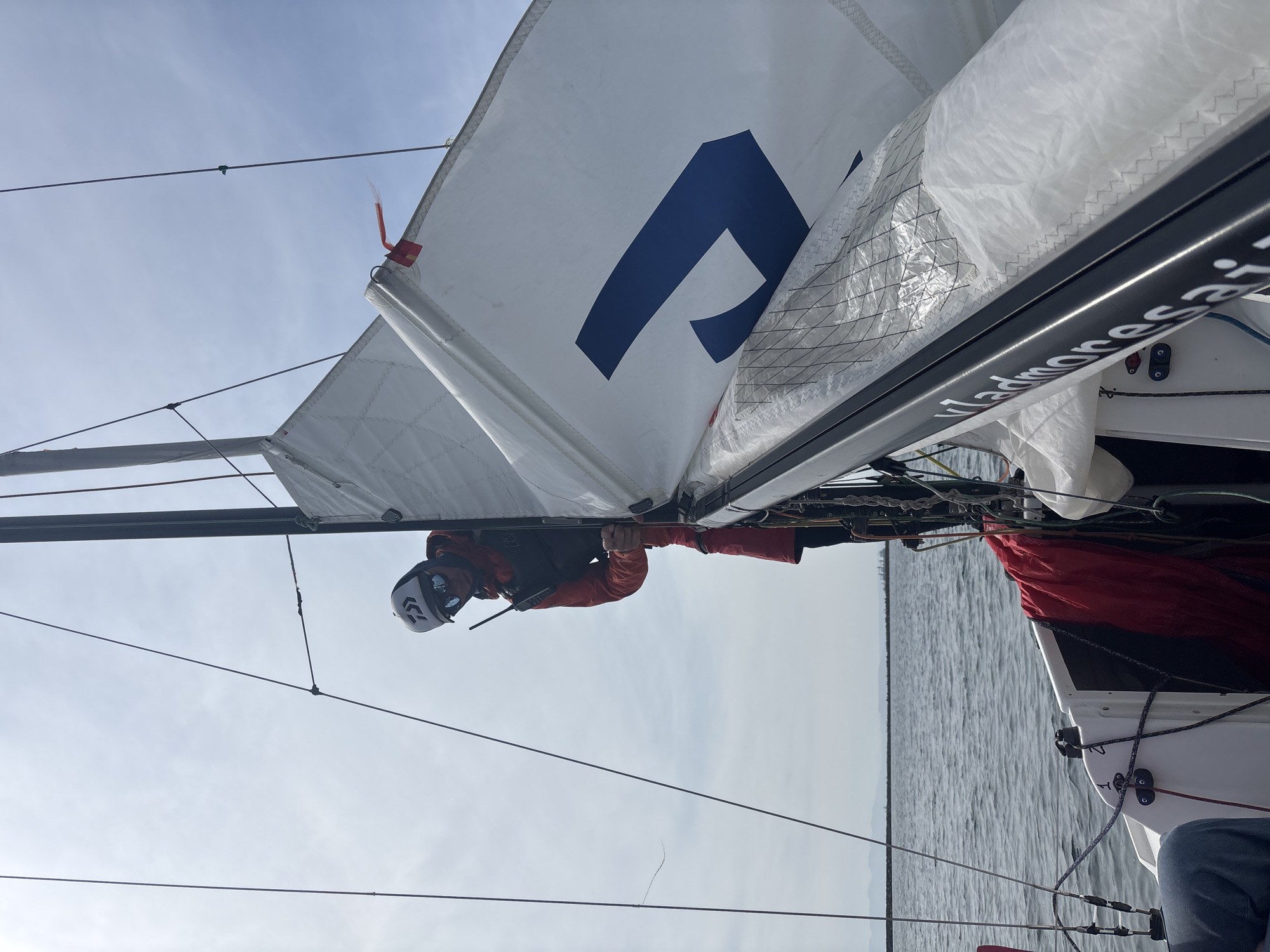 A person is on a sailboat. The sailboat has a white sail with a blue logo on it. The person is wearing a helmet, orange jacket, and black life vest. The person is holding on to the boom. The boom has the word "Vladmore" on it. The water is calm. The sky is cloudy. The location is not in the city of Vladivostok, Russia.