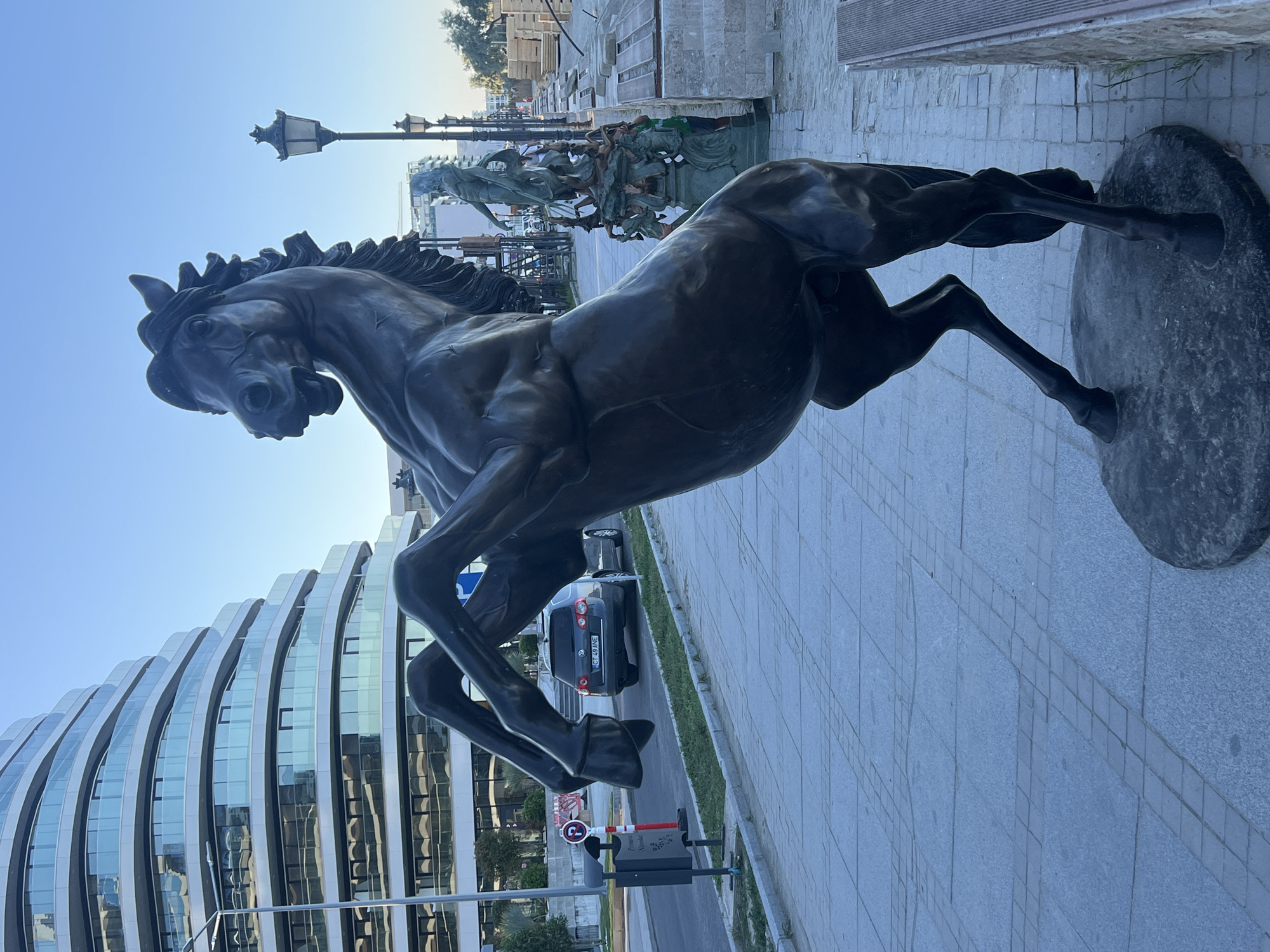 A bronze statue of a rearing horse stands on a stone base on a paved walkway. The horse is dark brown and detailed, with its front legs raised in the air and its head turned to the left. In the background, a modern building with curved glass balconies is visible. To the right of the statue, there is a street with a car and a traffic sign. Further in the background, there is a lamp post and another statue of a person. The scene appears to be in a public area in the city of Năvodari, Romania, likely a park or promenade, with construction materials visible in the far background. The sky is clear and blue, suggesting it is daytime.