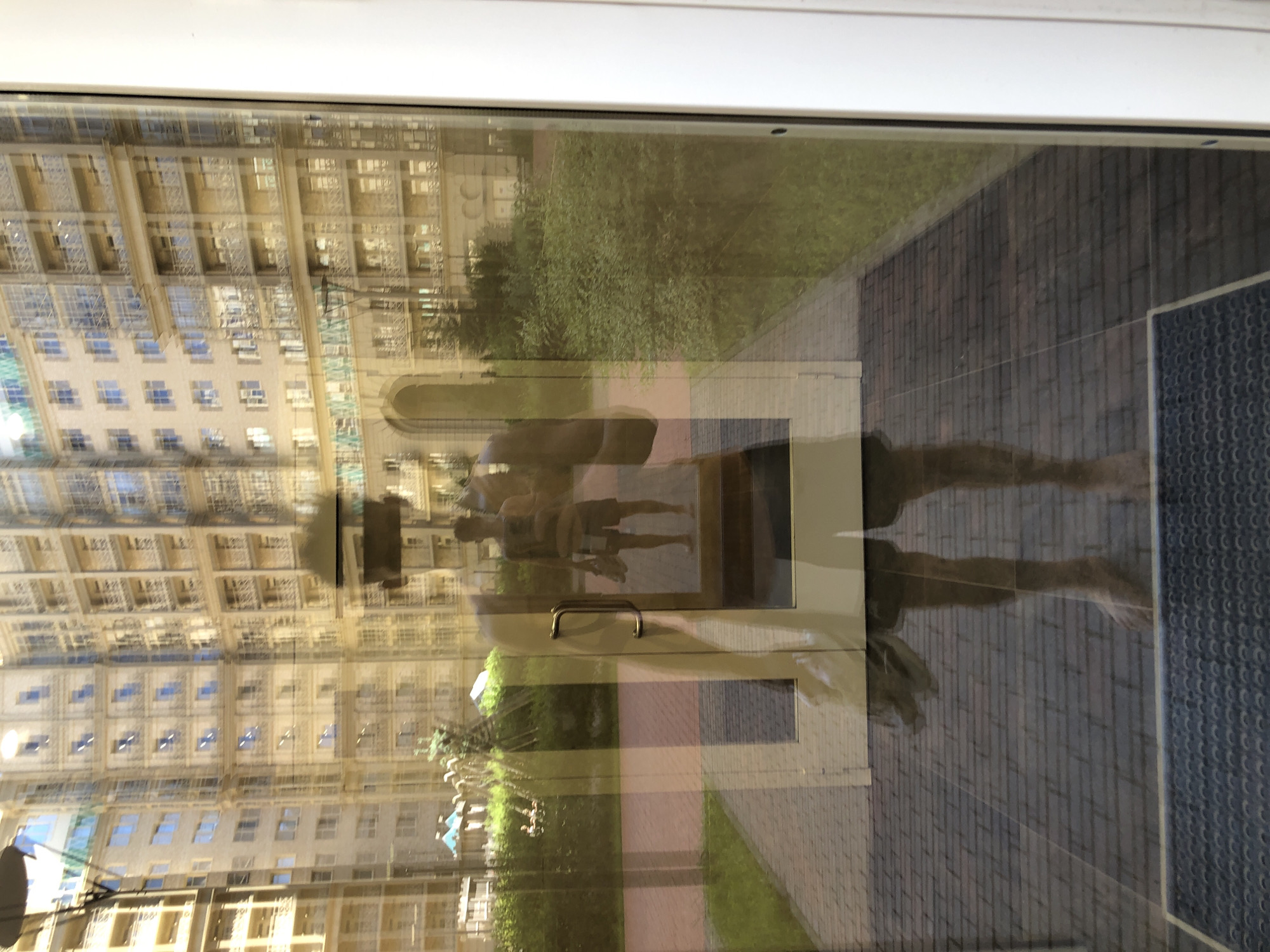 This image captures a person's reflection in a large glass door or window, looking out onto a modern residential complex in Saint Petersburg, Russia.
The foreground is dominated by the reflection of a male figure, seen from mid-thigh up. He is wearing a dark, sleeveless top and dark shorts. He holds a dark smartphone in his right hand, angled towards the glass, indicating he is taking the photograph. His face is obscured by shadow and the phone's position; his feet are bare. A metallic door handle is prominently visible on the right side of the glass panel, confirming it as a door. The very bottom of the image shows a dark grey, patterned mat on the floor inside, partially covered by the reflection of the person's feet.
Through the glass, and integrated with the reflection, an exterior scene is visible. A large, multi-story residential building complex with light-colored facades forms the background. It features numerous windows, balconies with railings, and architectural details such as arches and cornices, characteristic of modern or neoclassical revival construction. A paved pathway, made of interlocking grey and reddish-brown bricks, leads away from the door. Areas of green grass and dense shrubs line the pathway and fill parts of the courtyard. In the mid-ground, several small structures with distinctive blue, conical roofs can be observed, possibly play equipment or small gazebos. The scene is brightly lit by natural daylight, contributing to the strong reflections on the glass surface.