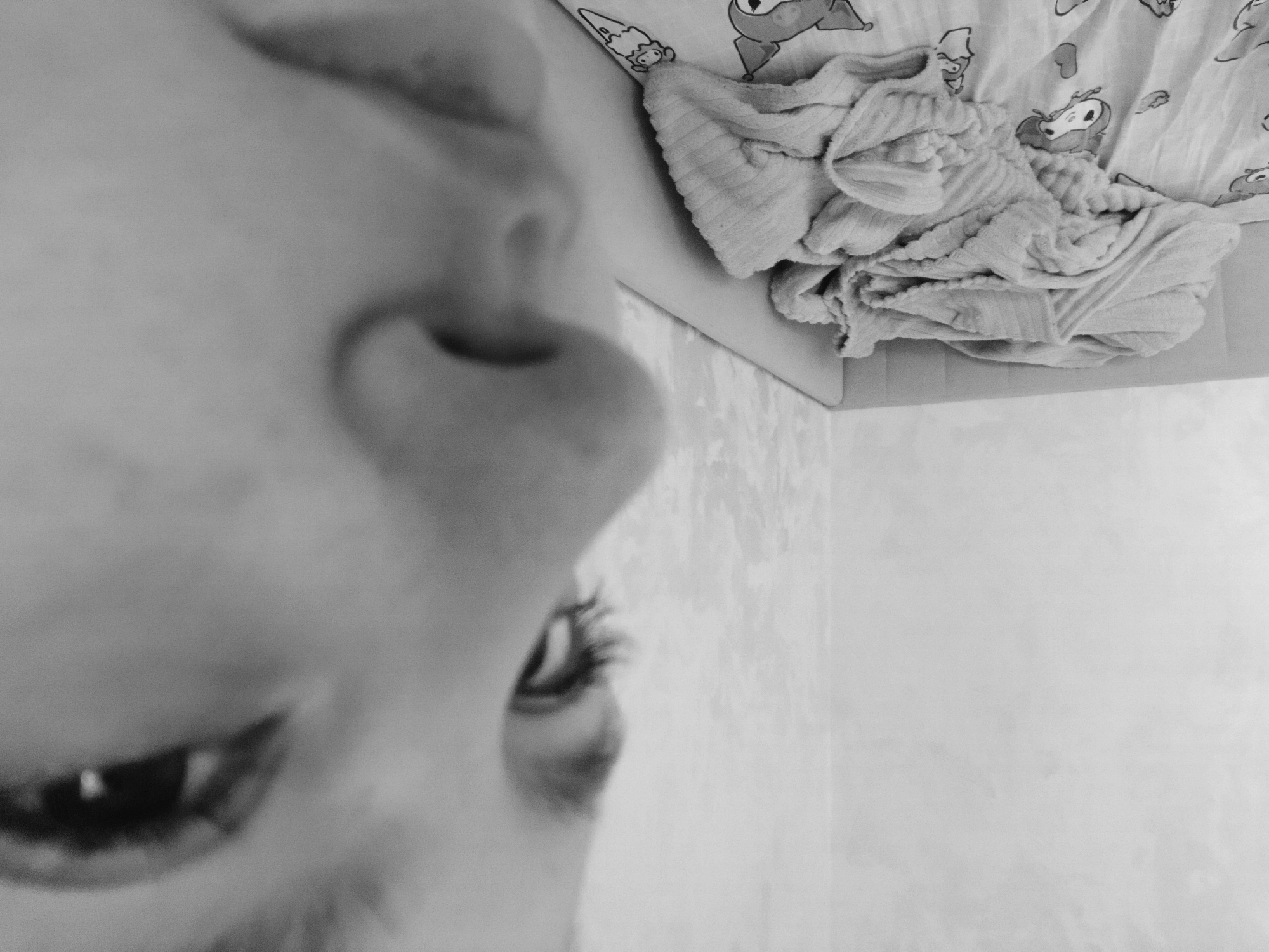 The image is a close-up, tilted view of a person's face, specifically the nose, one eye with eyelashes, and part of the mouth. The background includes a wall with a subtle texture and a cardboard box containing a folded, light-colored blanket with cartoon designs on the top sheet. The location, Марковский, Russia, is not identifiable in the image.