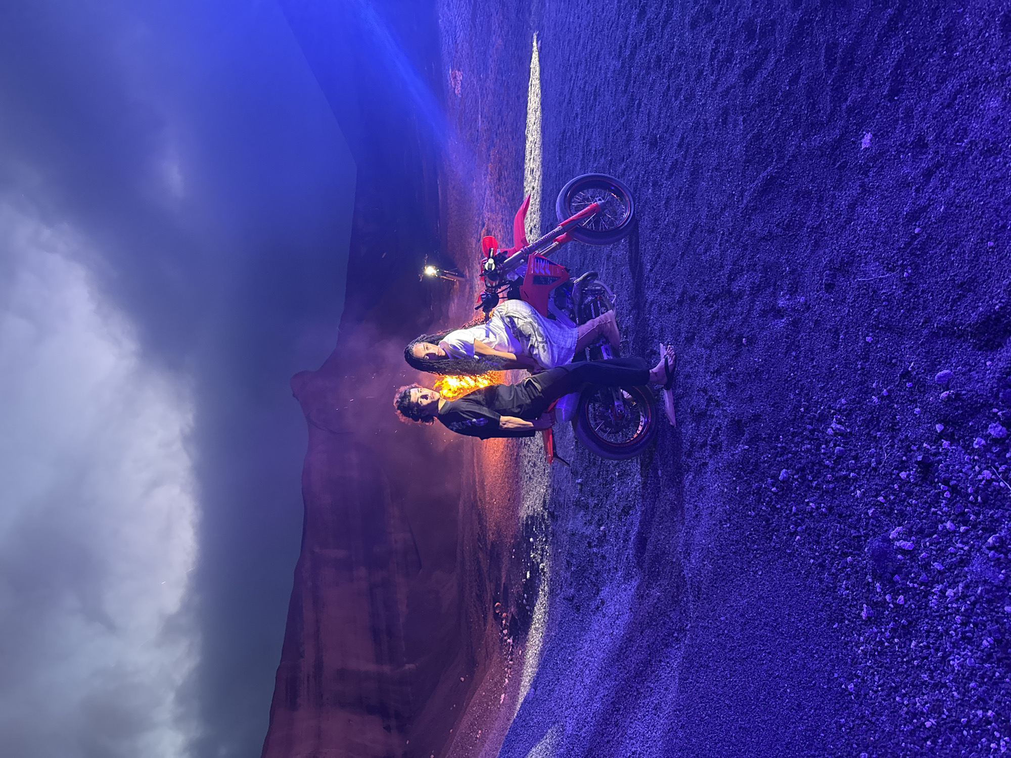 The image shows two individuals posing with a red dirt bike on a gravel surface under blue lighting. Behind them is a scene that resembles a canyon or rock formation. The sky is partially visible, with clouds. There is a small light source visible in the background, near the canyon. The location is likely near a volcanic area and possibly an arena, which suggests that it could be in Teguise, Spain. However, without more context or information, the exact location is hard to pinpoint.