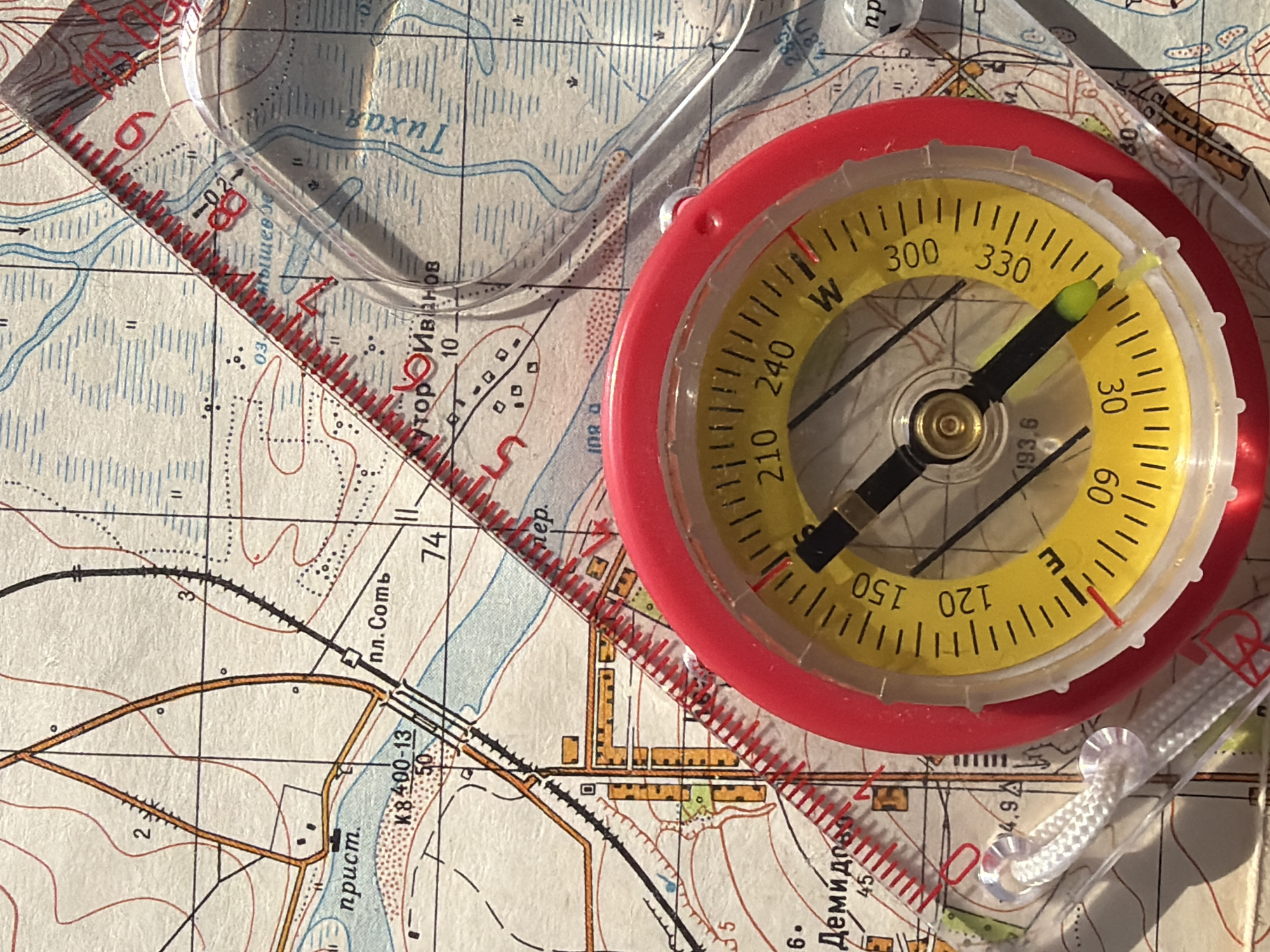 The image shows a compass and a ruler placed on a map, possibly for navigation purposes. The compass has a red rim and a yellow face with black lettering indicating directions (E, W) and degree markings. The needle points towards approximately 30 degrees. The ruler is clear with red markings along its edge. The map underneath features terrain lines, gridlines, roads, and other geographical information printed in various colors including blue, brown, and red. The text on the map is in Cyrillic. The city location seems to be near Демидов, пл. Соть, Тихая, and тор Иванов, possibly indicating a location in or around Izhevsk, Russia. There is a railroad shown.
