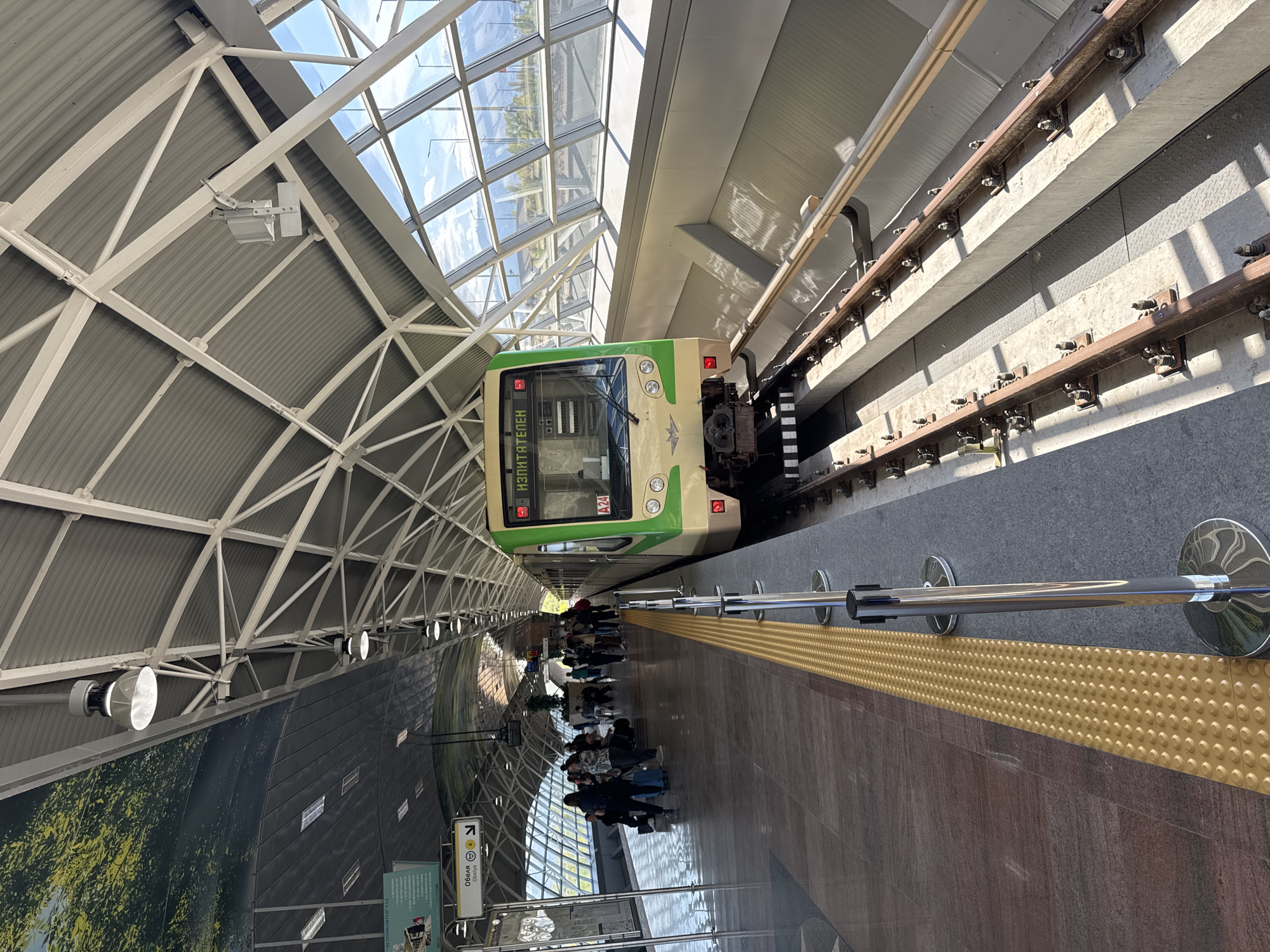 This image captures a vibrant, modern metro station in Sofia, Bulgaria, bathed in natural light. A green and beige metro train, marked with "ИЗПИТАТЕЛЕН" (IZPITATELEN), which translates to "TEST," and the code "A24" on its front, is stopped at the platform. The train's front features headlights and a dark windshield.
The station boasts impressive architecture, with a high, vaulted ceiling composed of white metal trusses, corrugated panels, and extensive glass skylights that reveal a bright, clear sky. This design allows abundant sunlight to illuminate the platform.
On the platform, a group of people, appearing to be passengers, are sitting and standing, possibly waiting for the train or having just arrived. The platform itself has a polished, reflective dark floor, alongside a prominent yellow tactile paving strip designed for the visually impaired. A chrome safety railing runs parallel to the track. On the wall opposite the train, a large mural depicting green foliage adds a natural element to the urban environment. A white directional sign with a left-pointing arrow clearly indicates "Обеля" (Obelya), a metro station in Sofia, with its English transliteration below. The metro tracks, securely fastened to concrete structures, are visible to the right of the train. The scene depicts an active, daytime setting within a well-maintained and efficiently designed public transport space.