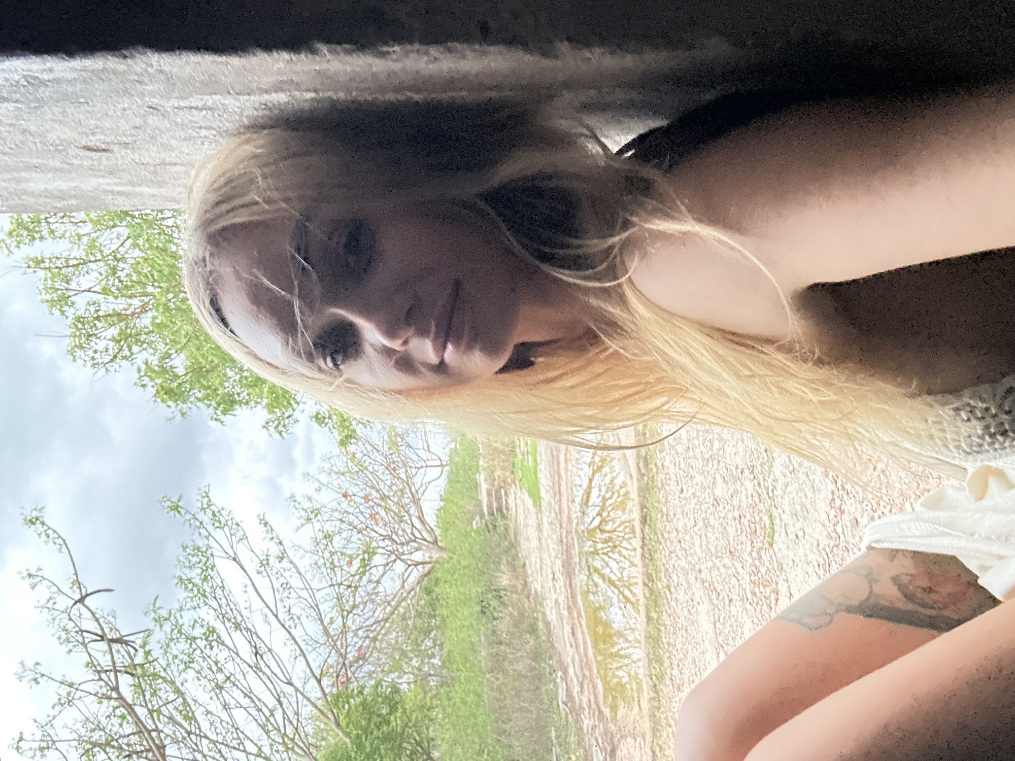 The image shows a woman with blonde hair, possibly sitting or leaning, with a view of a landscape in the background. The location appears to be Phu Thuy Ward, Vietnam, based on the presence of characteristic trees and vegetation in the background. The woman has a tattoo on her thigh and is wearing a light-colored garment. The background includes a dirt path, trees, and a cloudy sky. The image seems to be taken from a low angle, possibly from under a structure or shelter.