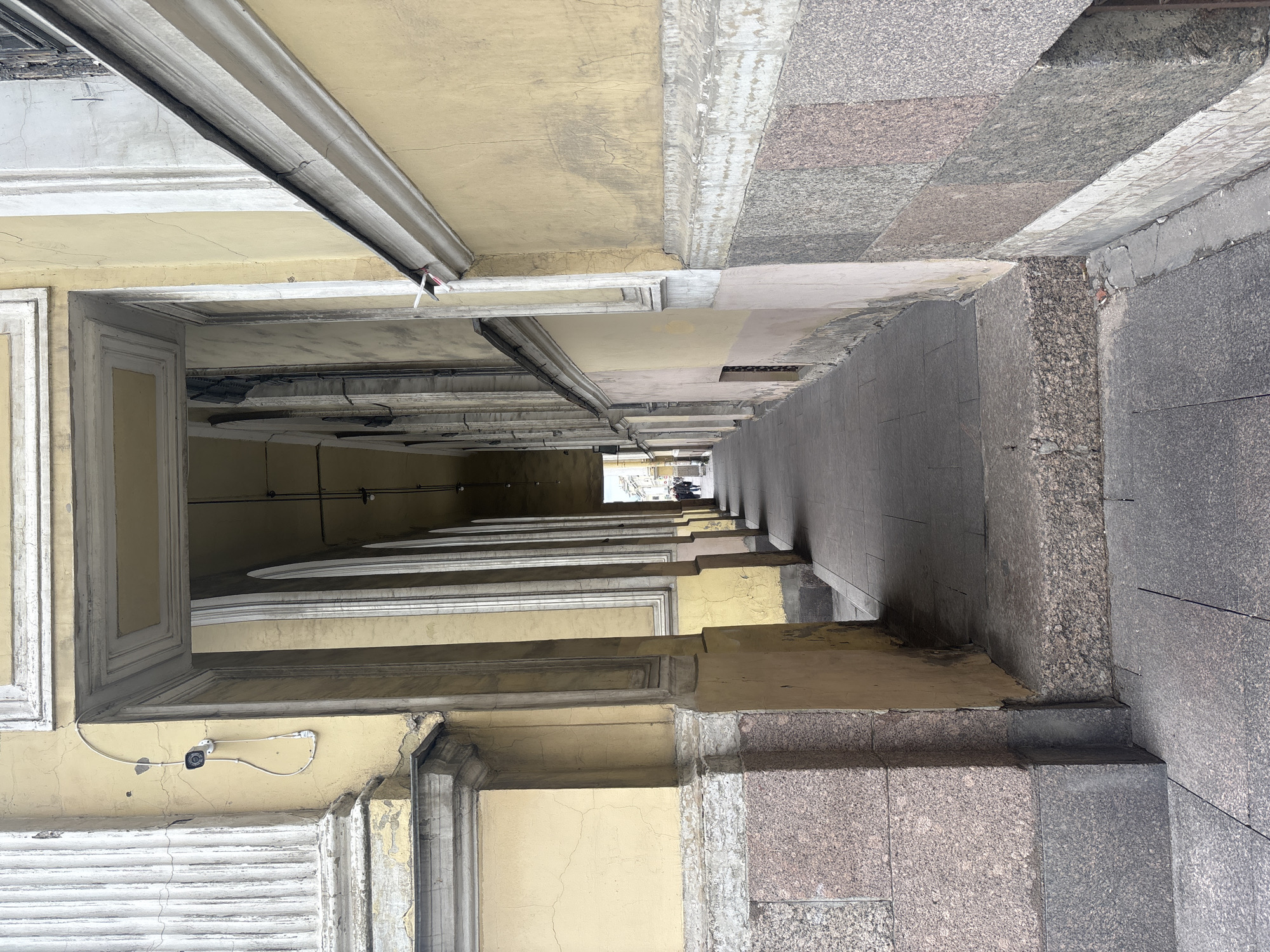 This photo depicts a narrow, covered alleyway in Saint Petersburg, Russia. The alleyway is characterized by aged, yellow buildings with classical architectural details lining either side. The buildings feature framed openings with decorative molding and, in one instance, a security camera attached to the exterior. The ground is paved with a mix of gray and reddish-brown stone slabs. At the far end of the alleyway, there is an opening to daylight, where several people appear to be standing, their figures obscured by the distance and contrast. The perspective creates a strong vanishing point, emphasizing the alleyway's length and narrowness.