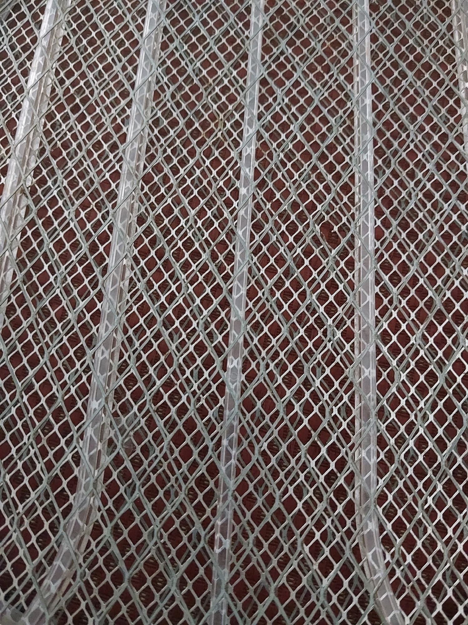 The image shows a close-up, overhead view of a light grey diamond-patterned mesh material. Behind the mesh, a darker reddish-brown surface is visible. The mesh is interspersed with several clear, cylindrical tubes or pipes running vertically through the frame. The lighting suggests an indoor or shaded outdoor environment. The image does not provide any contextual information about the city of Malang, Indonesia, or any specific details about people, actions, or interactions.