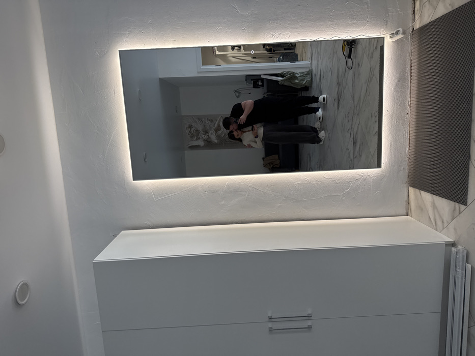 The image displays an interior room, stated to be located in Chaikovsky, Russia. A large, rectangular mirror with integrated perimeter backlighting is centrally positioned against a textured white wall.
Reflected in the mirror are two individuals: a female with dark hair, wearing a light-colored long-sleeved top, dark trousers, and white sneakers, holding a smartphone; and a male with a beard, dressed in a black t-shirt, black trousers, and white slides. The male's left arm is around the female's left shoulder, and he holds a black smartphone, appearing to take a mirror photograph of himself and the female. The reflected background behind them features a white bas-relief sculpture depicting winged figures on a wall, a dark sofa draped with fabric, and an open doorway leading to another room containing shelves and miscellaneous items.
Outside the mirror's reflection, to the left, stands a tall, white two-door wardrobe with two vertical silver handles. Several white trim pieces are stacked horizontally on the light-colored tiled floor beneath the wardrobe. To the right of the mirror, a white double electrical outlet is visible near the floor, with a black power cord extending from it to a yellow power tool resting on the floor. A grey, textured rectangular mat lies on the tiled floor directly in front of the mirror. The white ceiling contains two recessed light fixtures.