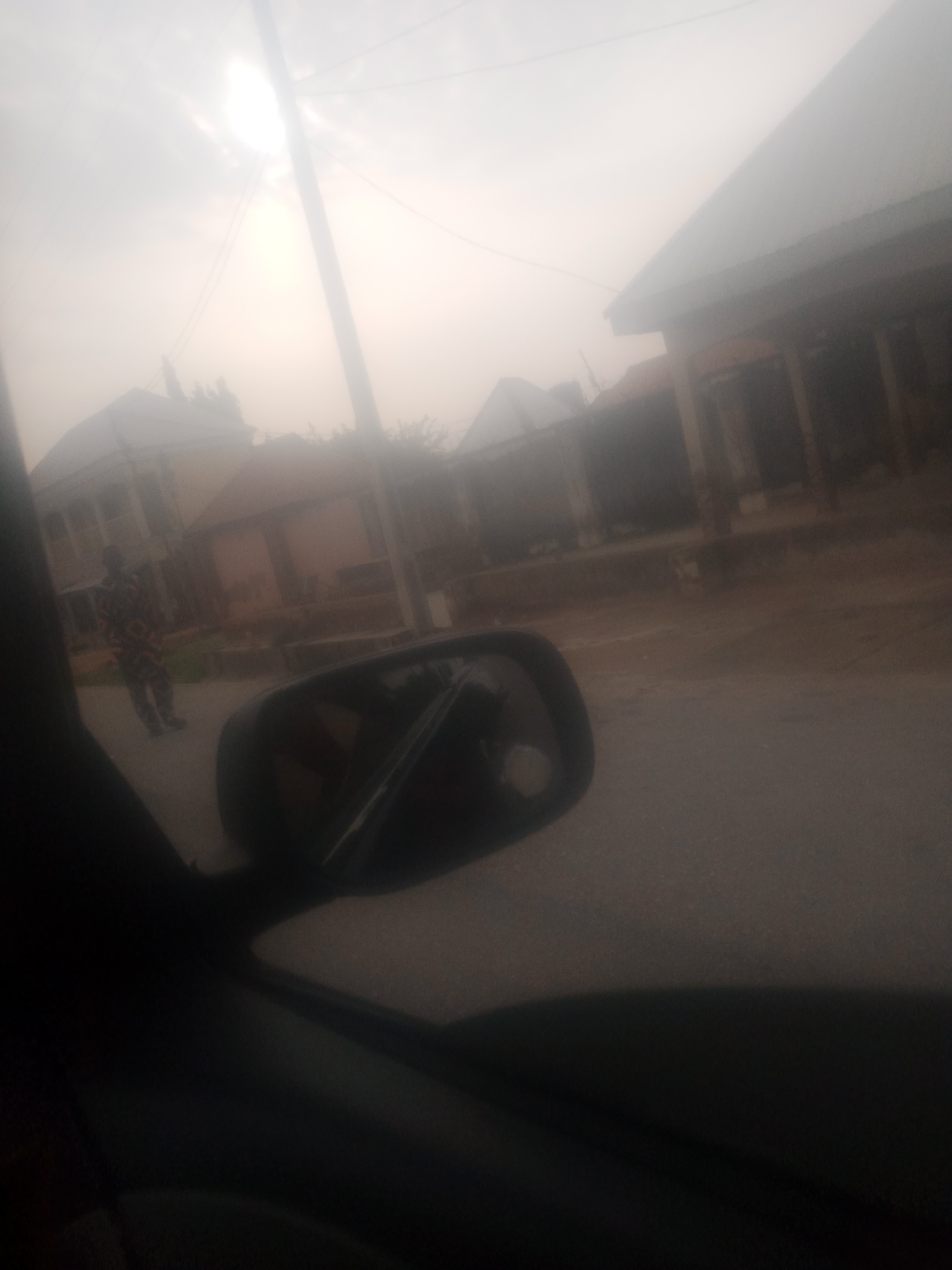 The image is a blurry, overcast view from inside a vehicle, looking out towards a street in Jalingo, Nigeria. A person in a distinctive patterned outfit, possibly a uniform, stands on the sidewalk to the left. In the background, there are buildings with corrugated metal roofs and some vegetation. A utility pole and power lines are visible in the sky. The overall impression is of a somewhat hazy and subdued urban environment.