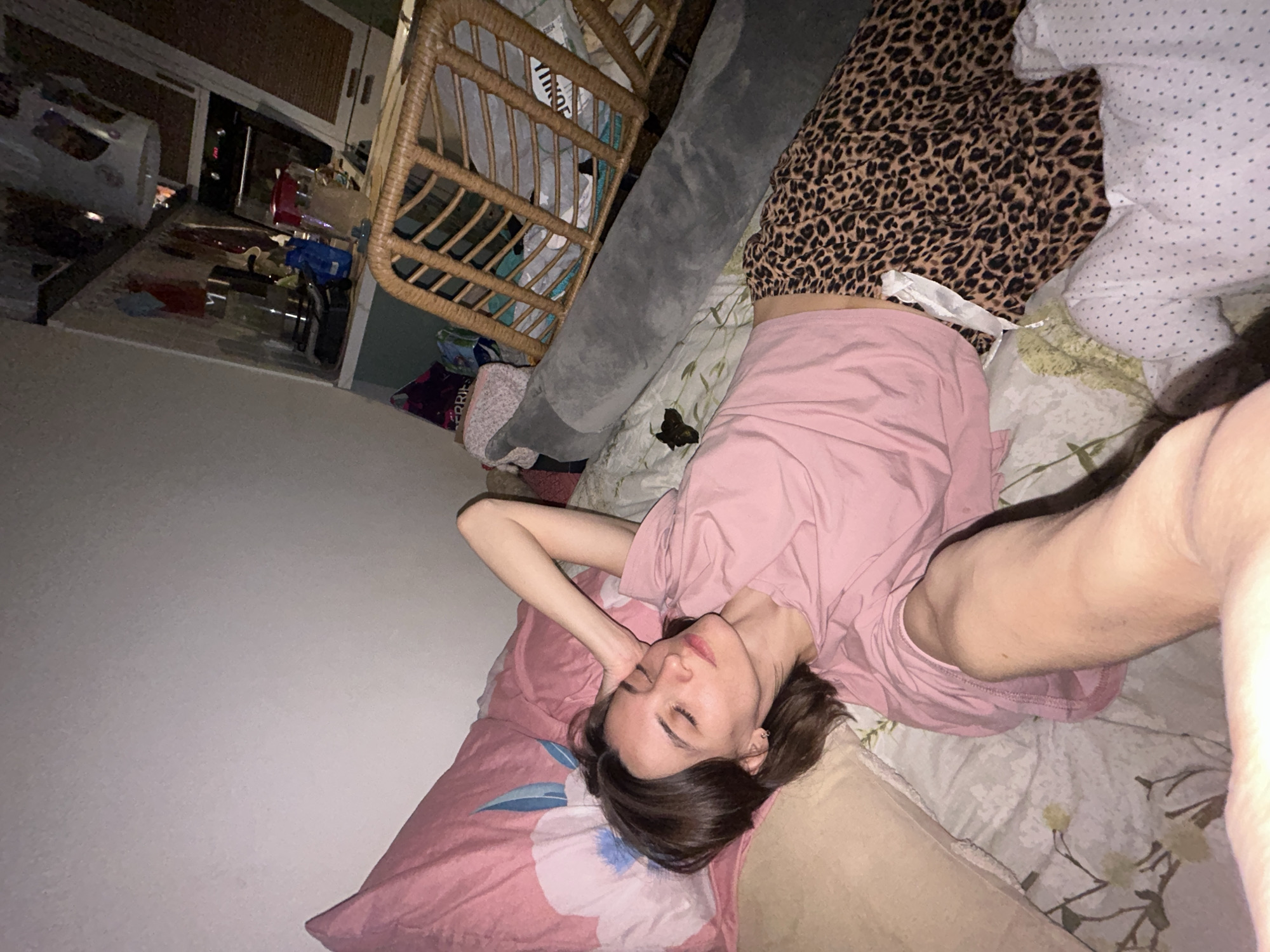 A person is lying on a bed with a pink floral pillow in a room. The person is wearing a pink shirt. The bed has patterned sheets, including a white dotted pattern and a leopard print pattern. Beside the bed is a wicker storage unit, a grey body pillow, and various cluttered items. The location appears to be indoors and possibly a domestic setting, but there is no information present in the image to confirm it is Moscow, Russia.