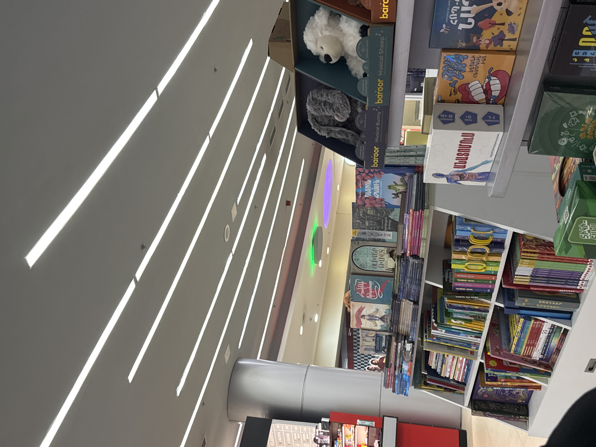 The image captures an interior view of a commercial establishment, likely a bookstore or a toy store, in Yerevan, Armenia. The perspective is tilted, showcasing a high, modern ceiling with numerous long, bright linear light fixtures arranged parallel to each other across a gray surface.
On the right side of the frame, several white shelving units are packed with various products. An upper shelf displays two boxed plush toys: a gray "baroor Musical Bunny" and a white "baroor Musical Sheep." Below these, numerous books and boxed games are neatly arranged, some stacked vertically, others horizontally. Many book covers feature colorful illustrations and titles predominantly in Armenian script.
Visible Armenian titles include "Ո՞վ կերավ Բալուս" (Who ate Balus?), "Կլին-Կլա," "Պահպանեք Ընկերոջը" (Protect a Friend), "Հրաշքներ աշխարհում" (Wonders of the World), and "Իրականը" (The Real One). One prominent white box, possibly a board game, is titled "ՊԱՏՈՒՍՏՈՒՄ" (Patutsum) and depicts a human anatomy illustration, along with age ratings like "12+" and "30-60." Another book cover partially reveals the English text "WHY EARTH MAN..."
In the mid-ground, a large, silver-colored cylindrical pillar extends from the floor. Beyond the shelving, glimpses of a broader retail space are visible, accented by subtle colored ambient lighting in hues of purple and green. The scene suggests a well-lit and organized retail environment, focusing on educational items, books, and toys. No distinct individuals are clearly visible.