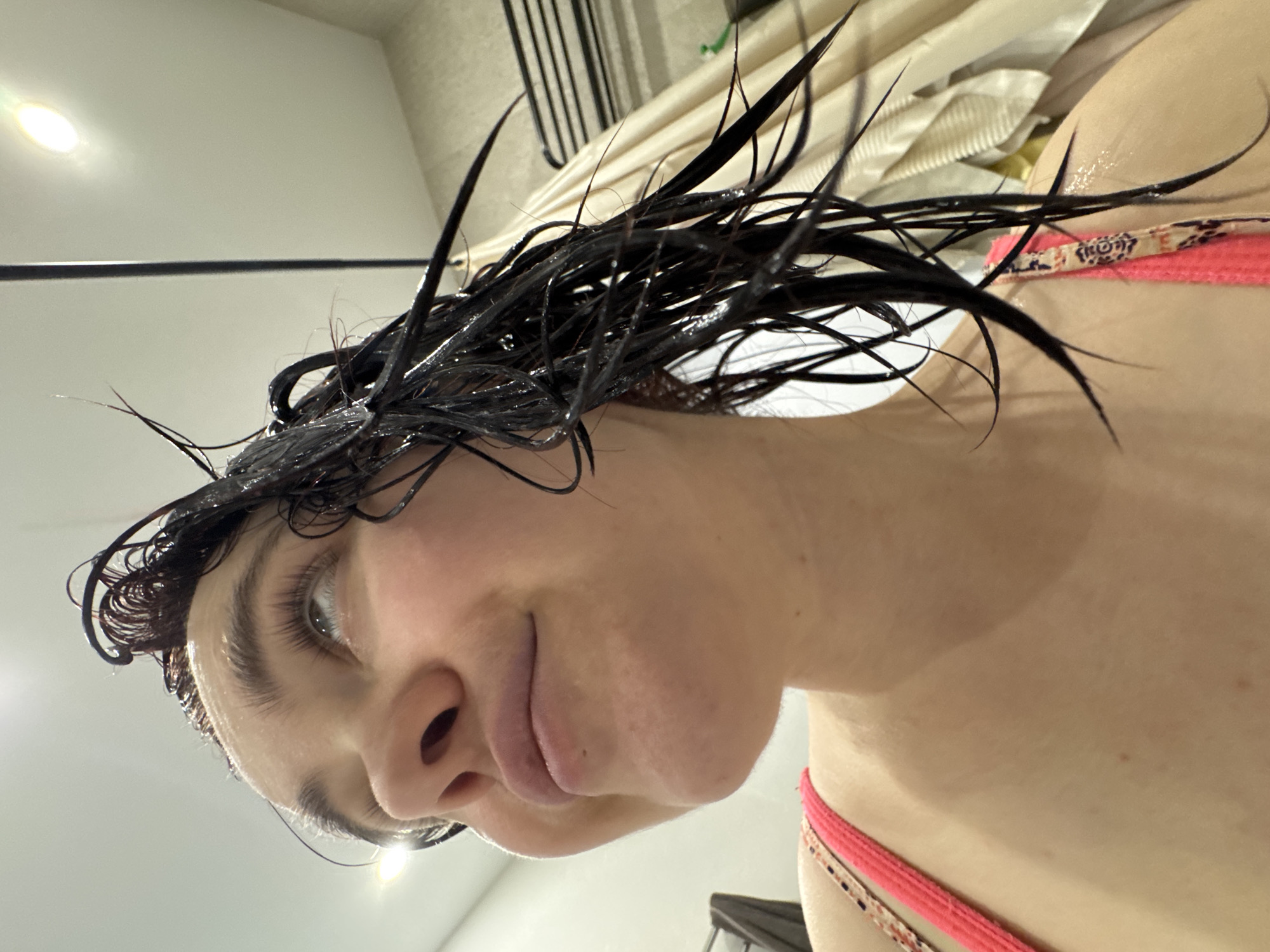 A close-up, low-angle shot shows a person's face and upper body against a blurred background suggesting a bathroom. The person has wet, dark hair sticking up and out in multiple directions. Their skin is fair. They are wearing a swimsuit top with a pink strap. The background includes a shower curtain and what appears to be a towel rack. There is no indication that the location is Izhevsk, Russia.