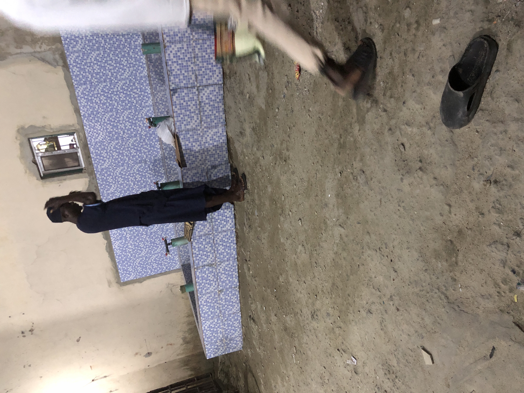 The image depicts an outdoor scene in Ado, Nigeria, possibly near a public facility. In the foreground, a dark-colored sandal lies on the dirt ground. Further back, a person in light-colored pants is blurred, suggesting movement. To the left, a person wearing a dark blue outfit and a cap stands with their hands on their head, facing a wall adorned with blue and white checkered tiles. This tiled wall appears to be part of a bathing or washing area, with several green spigots visible. A small, barred window is set into the wall above the tiled area. The ground is uneven and covered with dirt and scattered debris.