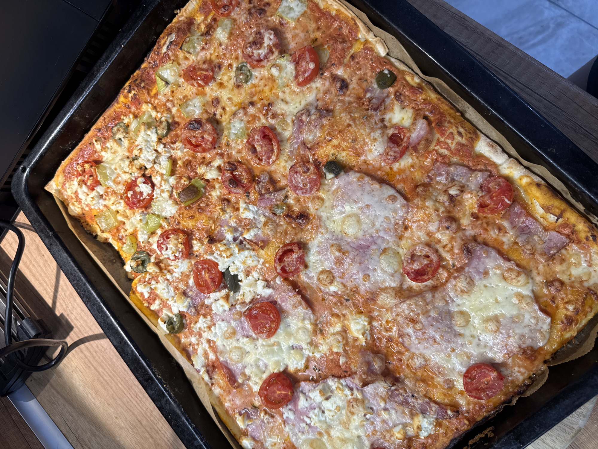 A rectangular pizza is shown in a black baking tray, placed on a wooden surface. The pizza is topped with tomato sauce, cheese, ham, cherry tomatoes, feta cheese, and green peppers. The scene appears to be indoors, possibly in a kitchen. There is no discernible information within the image to suggest a specific location in the city of Varna, Bulgaria.