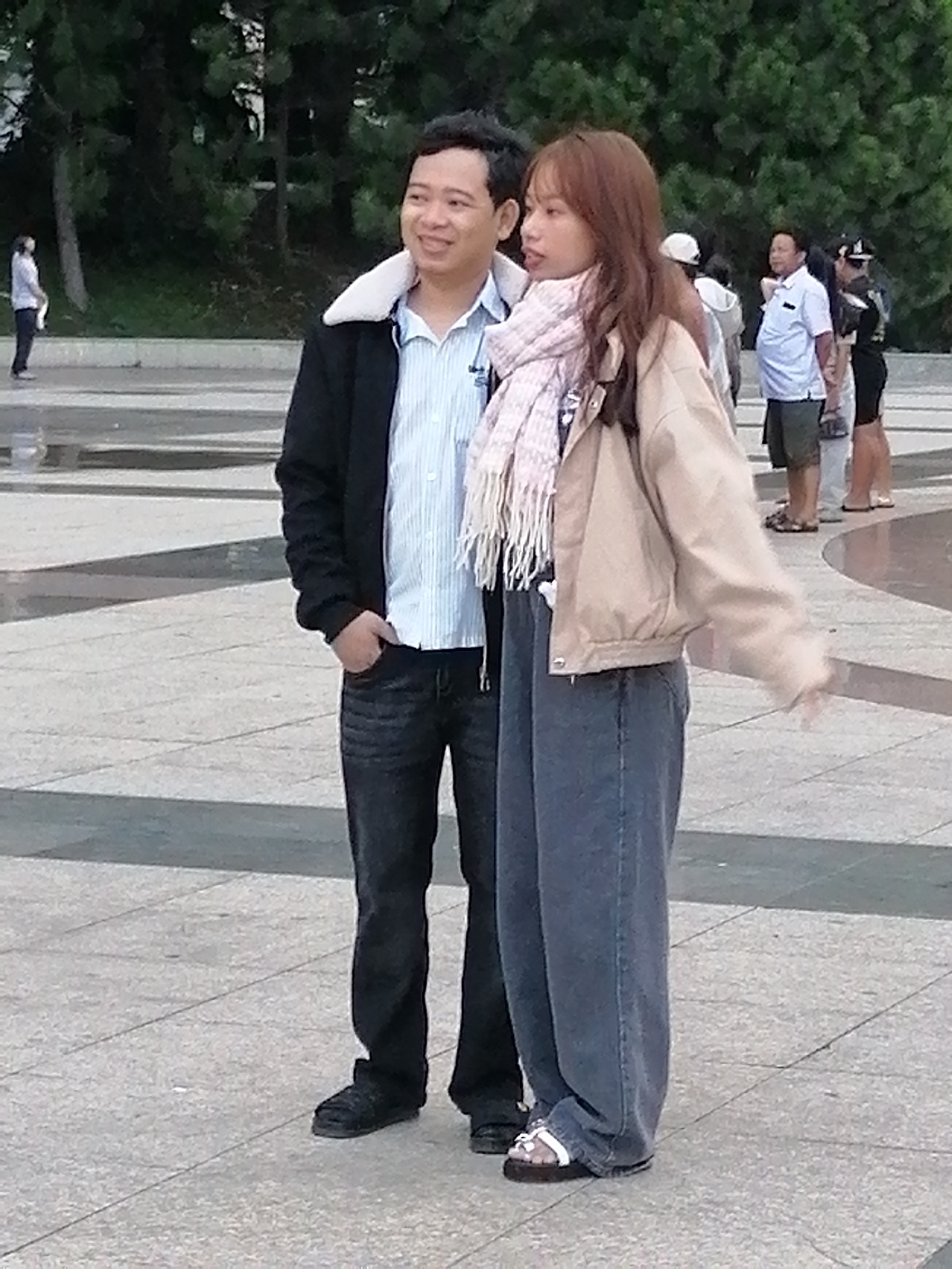 The image captures two individuals, a man and a woman, standing on a paved outdoor area with trees in the background. The man, on the left, is wearing a black jacket with a white collar, a light blue striped shirt, and dark jeans. He has his right hand in his pocket. The woman, on the right, is wearing a light brown jacket over a patterned top and dark, loose-fitting pants. She has a light pink scarf around her neck. She is wearing white sandals. Behind them, other people are visible in the background, including a person in a white shirt and shorts, and another in a black shirt and shorts. The ground is paved with square tiles and has some patterned sections. The setting appears to be a public space in Da Lat, Vietnam, likely a park or promenade given the presence of trees and paved walkways. The overall lighting suggests it is daytime.