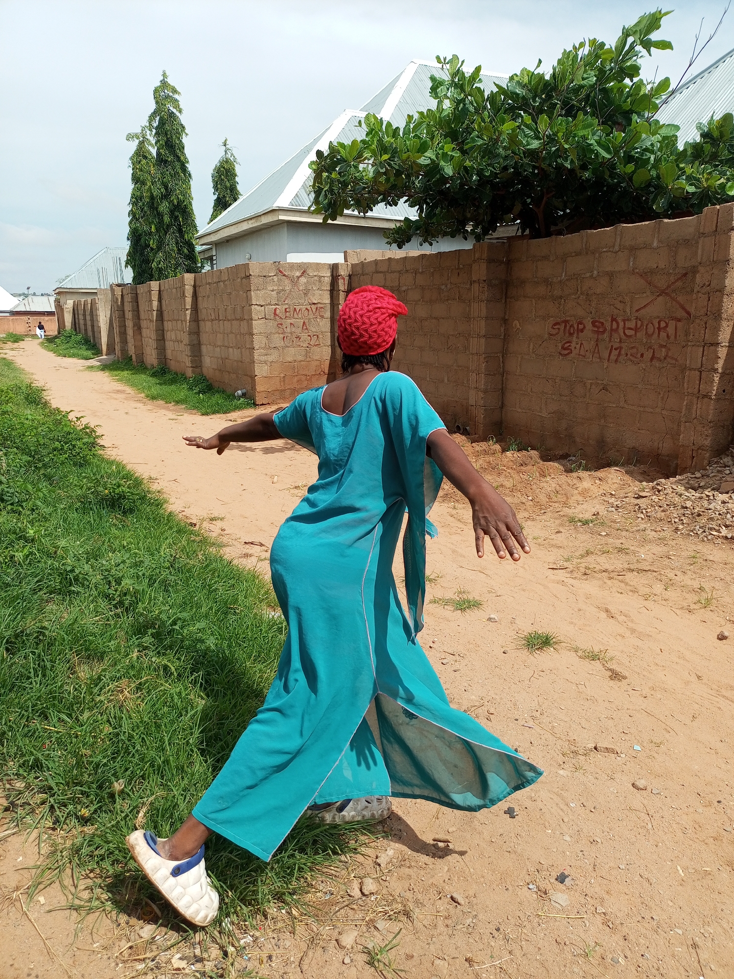 A Black woman wearing a teal dress and a red headscarf is shown from the back, with her left arm extended. She is standing on a dirt path lined with grass on the left and a brick wall on the right. The wall has red graffiti on it that reads "STOP REPORT S.L.A 17/3/23". In the background, there are houses with metal roofs and tall trees. The scene appears to be in a rural or suburban area.