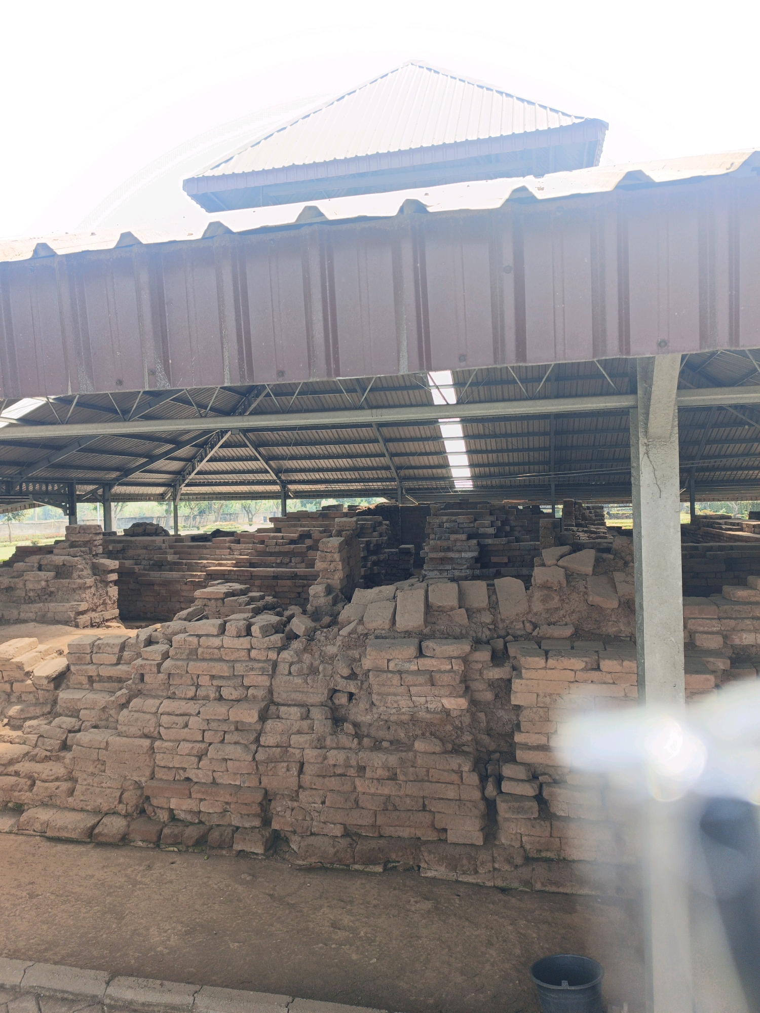 The image shows the archaeological ruins of an ancient structure, likely a temple or monument, in Kejagan, Indonesia. The ruins are made of brick and are partially excavated, with visible layers and foundations. A protective roof structure, made of metal sheeting and supported by metal beams and concrete pillars, covers the archaeological site to shield it from the elements. The environment appears to be outdoors, with a dirt ground and some vegetation visible in the background through the open sides of the covered structure. The lighting suggests it is daytime, with sunlight filtering through the roof. There are no people visible in the image. A dark bucket is placed on the ground near the right side of the frame.