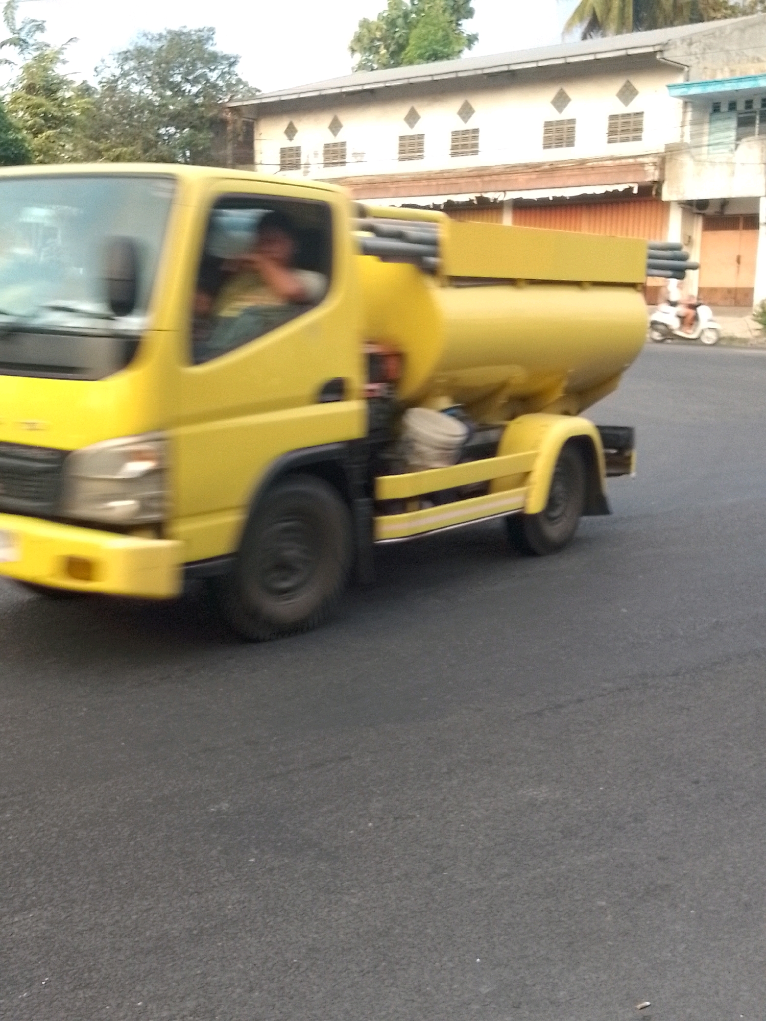 A yellow tanker truck is driving on a paved road. The truck has a large tank on its back with pipes extending from the top. The driver is visible in the cab, wearing a light-colored shirt. In the background, there are buildings with windows and awnings, and trees. A white scooter with a rider is also visible on the right side of the road. The image appears to be taken in Sukorambi, Indonesia, given the architectural style and vegetation.