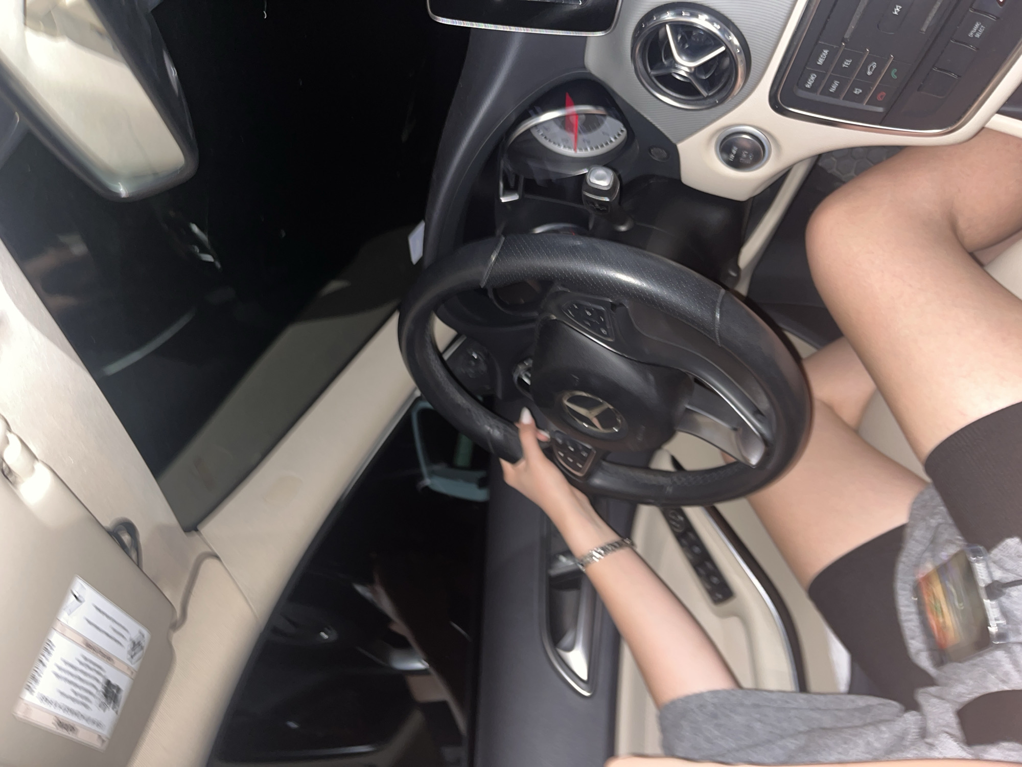 A person is seated in the driver's seat of a Mercedes-Benz car. The person is wearing grey clothing and black thigh-high socks. The person's left hand is on the steering wheel, with a silver bracelet visible on the left wrist. The car's dashboard, steering wheel, and interior are visible. The background outside the car windows is dark. The location is not possible to determine based on the image.
