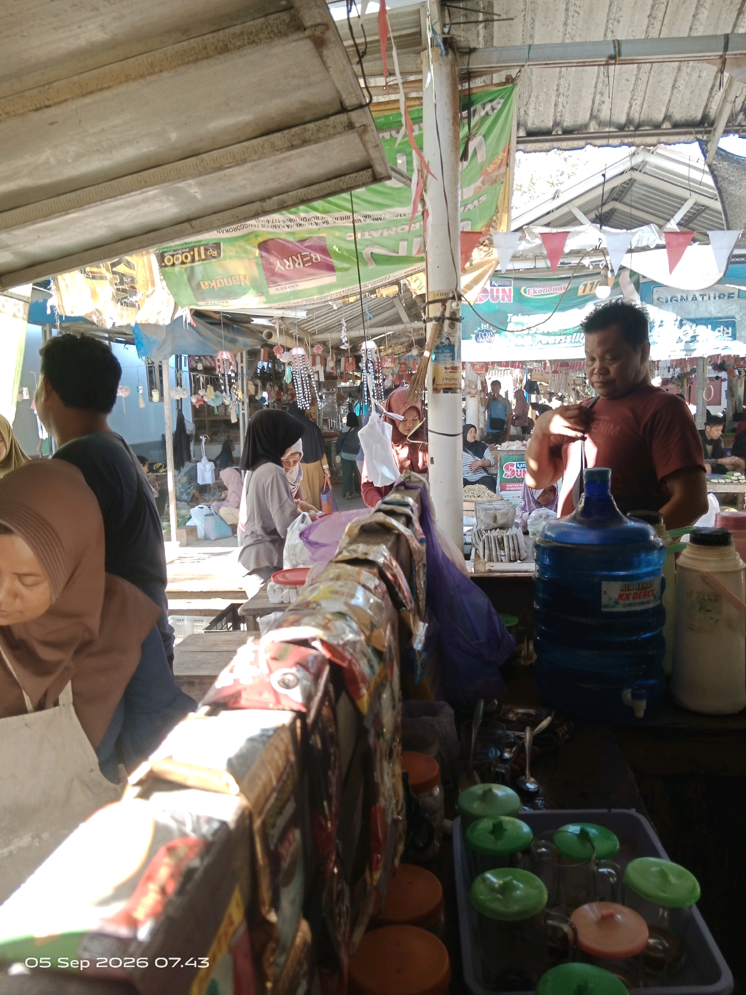 This is a medium shot, eye-level, outdoor photograph taken at a market in Kalipurwo, Indonesia. The scene depicts a busy marketplace with several individuals. In the foreground, on the right, a man in a red t-shirt is standing behind a large blue water dispenser. He appears to be serving or attending to customers. In front of the dispenser, a tray holds several glasses with colorful lids. On the left side of the image, partially obscured, are two women, one wearing a brown hijab and the other a darker hijab, both appearing to be customers. In the background, there are stalls with various goods, including what appear to be textiles and other merchandise. The market is covered by a corrugated metal roof, and there are banners and decorations visible. The lighting suggests it is daytime, with sunlight filtering through the structures. A timestamp at the bottom of the image reads "05 Sep 2026 07.43".