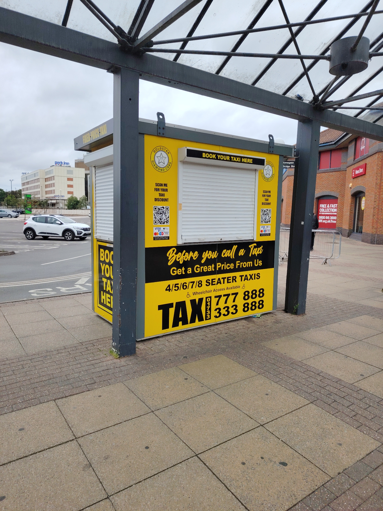 This image captures an outdoor scene in Peterborough, United Kingdom, featuring a bright yellow taxi booking kiosk positioned under a metal-framed glass canopy. The kiosk displays "BOOK YOUR TAXI HERE" and "FIXED-PRICE TAXI" on its top section. Its front panel features the "GOLDSTAR DIAMOND CARS" logo, two QR codes, and text reading "SCAN ME FOR YOUR TAXI DISCOUNT." Below this, "VISA," "Mastercard," "Apple Pay," and "Google Pay" logos are visible, indicating accepted payment methods. The lower section of the kiosk states "Before you call a Taxi Get a Great Price From Us," "4/5/6/7/8 SEATER TAXIS," and "Wheelchair Access Available." Two prominent phone numbers, "01733 777 888" and "333 888," are printed in large black text over a yellow background with "TAX" partially visible.
The kiosk stands on a paved pedestrian area composed of light-colored square concrete slabs and darker brick paving. To the left, a road or parking area is visible with a white SUV (Dacia Duster) moving or parked, and faint road markings on the asphalt. In the background, a multi-story building with "parkinn" signage is prominent, and an "ASDA" logo is visible on another distant building. To the right of the kiosk, part of a brick building with red window frames and a "Home Store" sign is present. A red sign behind the kiosk offers "FREE & FAST COLLECTION OF YOUR UNWANTED ITEMS 0800 915 3000 bnf.org.uk/free." The sky is overcast. No individuals are clearly depicted in the scene. Small green vegetation grows at the base of the kiosk's supporting pillar.