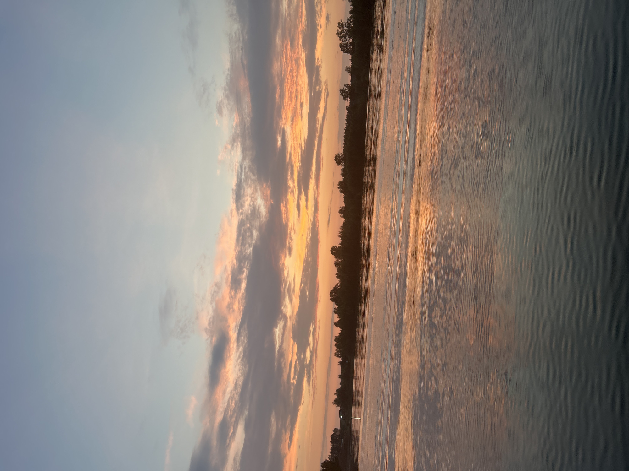 The image shows a wide river at sunset. The sky is filled with colorful clouds in hues of orange, pink, and purple, reflecting on the calm water. The opposite bank is lined with a dense forest. A few trees are visible on the horizon, and there appears to be a small building in the distance. The water's surface is smooth with gentle ripples, creating a mirror-like effect for the sky. The location is identified as Noviy, Russia, suggesting a riverfront area within or near the city.