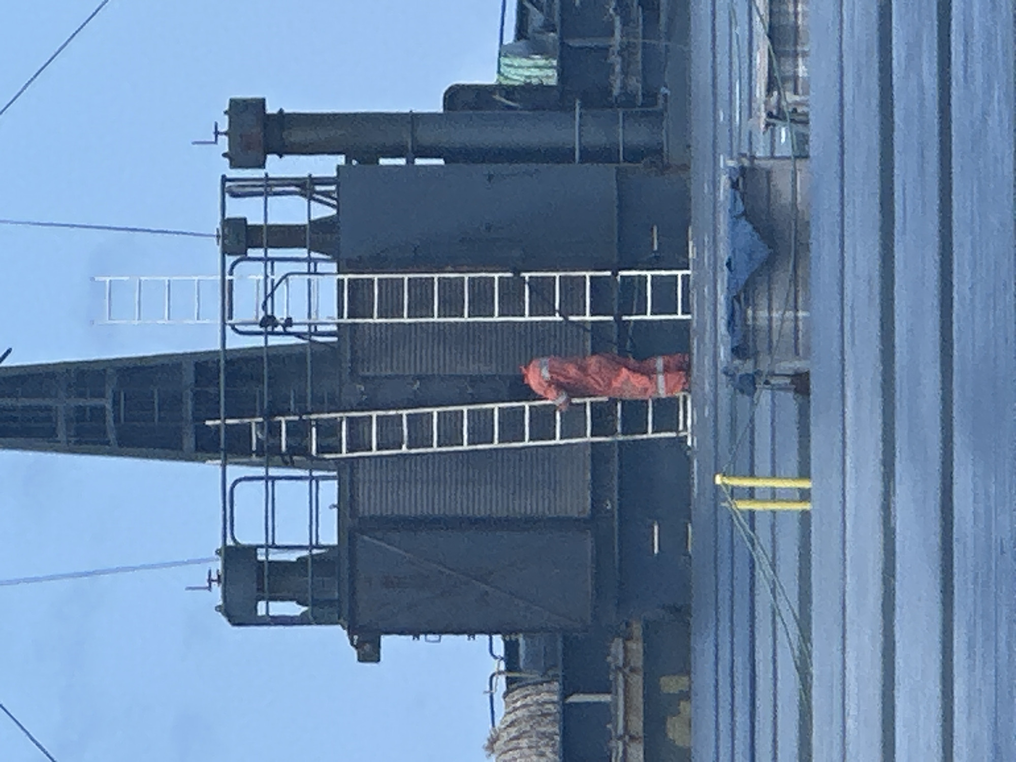 A medium shot depicts a piece of industrial equipment with a person climbing a ladder. The equipment appears to be some type of crane or hoist mechanism, featuring black metallic structures, multiple ladders, and a collection of cylindrical components at the top. The sky is visible in the background and the equipment is stationed on a ship. An individual clad in a bright orange outfit is seen ascending a ladder on the apparatus. Yellow supports are also visible. The location of the scene is not specified.