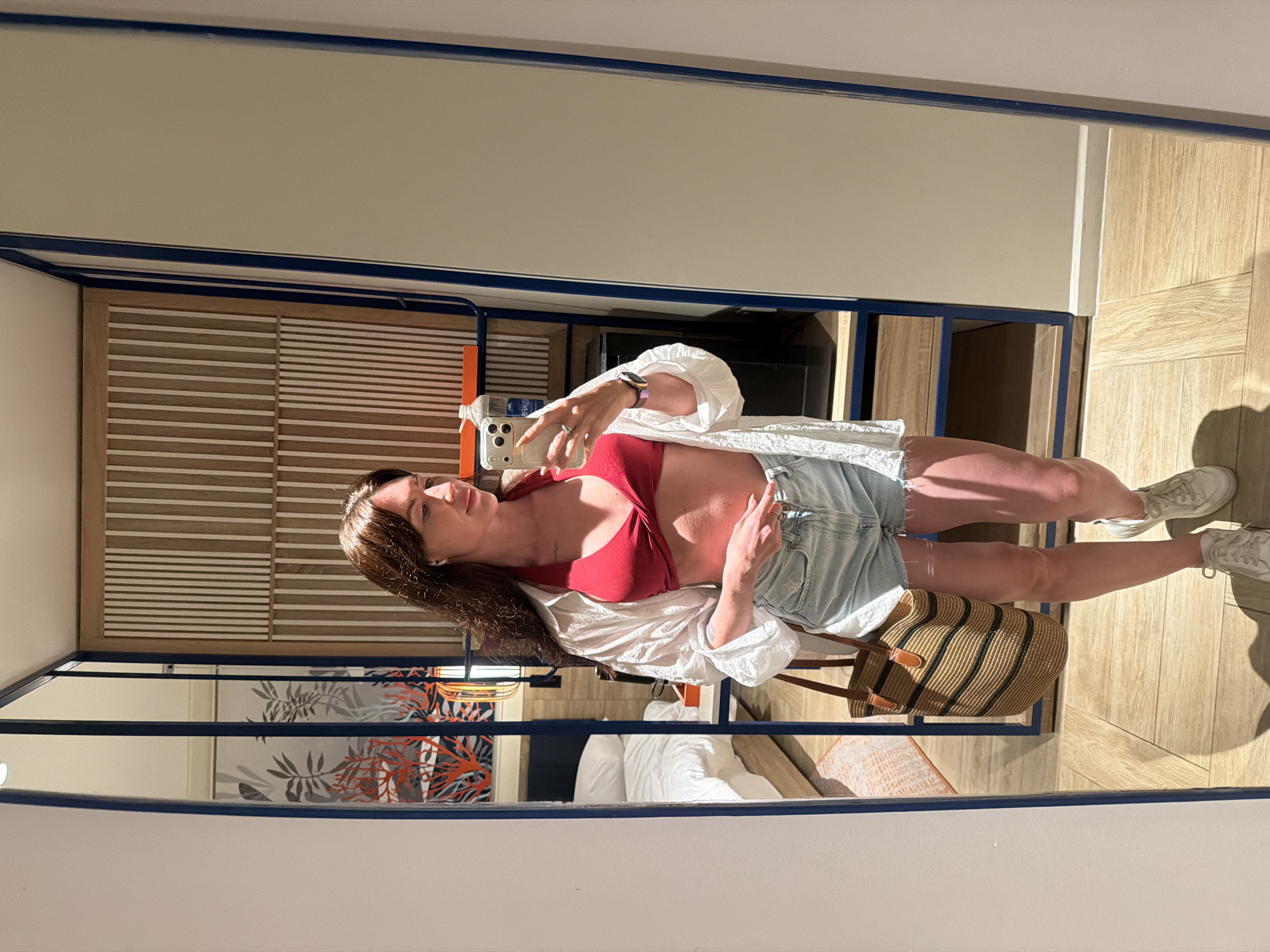 A woman is taking a mirror selfie in what appears to be a hotel room. She is wearing a red halter-neck bikini top, a white unbuttoned shirt, light-wash denim shorts, and white sneakers. She is holding a smartphone in her right hand and has a woven tote bag on her left side. The background shows a bed, a wooden accent wall, and part of a mirror frame. The image is taken in a well-lit indoor environment, likely a private accommodation, and there is no visible connection to Dong Ninh Hoa Ward, Vietnam.