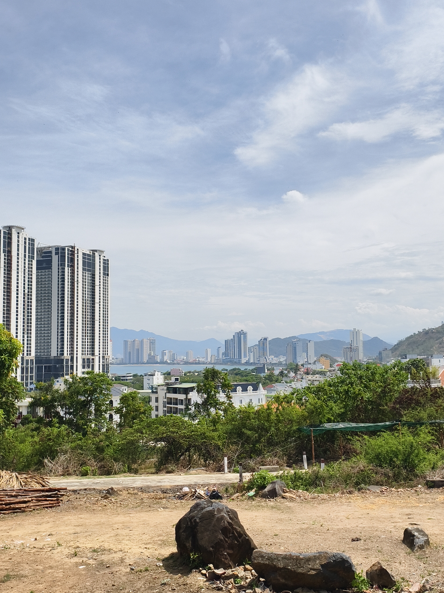 The image depicts a panoramic view in Bac Nha Trang Ward, Vietnam, likely from a partially developed elevated area. In the foreground, there's a rocky, dirt-covered ground with several medium-sized boulders and scattered debris. A pile of wooden logs is seen on the left side of the frame.
Midground vegetation consists of a mix of trees and shrubs, obscuring some residential structures beneath the prominent skyline. These structures include private houses with white rooftops as well as large apartment complexes.
The cityscape in the background features a cluster of tall buildings along a coastline, separated by water from distant mountainous terrain. The sky above is filled with light, scattered clouds. The overall impression is that the scene is captured during a sunny day.