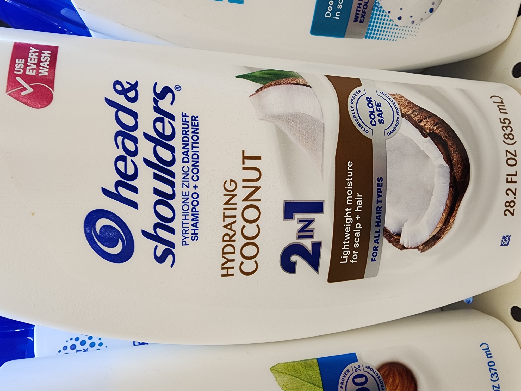 This is a close-up, eye-level, vertical, medium shot of a white plastic bottle of Head & Shoulders Hydrating Coconut 2-in-1 shampoo and conditioner. The bottle is positioned in the foreground and is partially obscured by another white bottle on its left and a blue-labeled bottle on its right. The bottle prominently displays the Head & Shoulders logo, which features a stylized eye within a circle, in blue. Below the logo, the text "head & shoulders" is written in a dark blue sans-serif font. Further down, "PYRITHIONE ZINC DANDRUFF SHAMPOO + CONDITIONER" is written in smaller, dark blue text. The primary descriptor, "HYDRATING COCONUT," is in large, dark brown, serif font. Below this, "2 IN 1" is displayed in large, blue, metallic-looking numbers and text. A graphic of a halved coconut with its flesh and husk is shown below the "2 IN 1" text, with a green leaf visible to the left of the coconut. A circular label with "CLINICALLY PROVEN COLOR SAFE DANDRUFF PROTECTION" is visible on the right side of the coconut graphic. A red checkmark icon with the text "USE EVERY WASH" is in the upper right corner of the bottle. The bottom of the bottle shows "28.2 FL OZ (835 mL)" and a small recycling symbol. The image was likely taken in a retail store environment, possibly a pharmacy or grocery store, given the presence of multiple product bottles and the shelving in the background. The specific location of Camp Hill, United States of America, is not discernible from the visual information in the image itself.