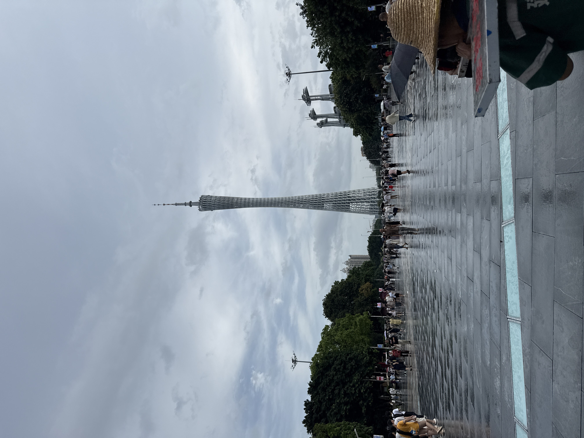 The image depicts a wide, paved pedestrian walkway in Tianhe District, China, with a prominent, slender observation tower in the background. The walkway is wet, suggesting recent rain, and lined with trees on either side. A crowd of people are walking along the pathway, many holding umbrellas, indicating continued or recent precipitation. In the foreground, a person wearing a straw hat is visible, facing away from the camera, with what appears to be a display board in front of them. The sky is overcast with a cloudy atmosphere. Several other modern structures and streetlights are scattered throughout the scene.