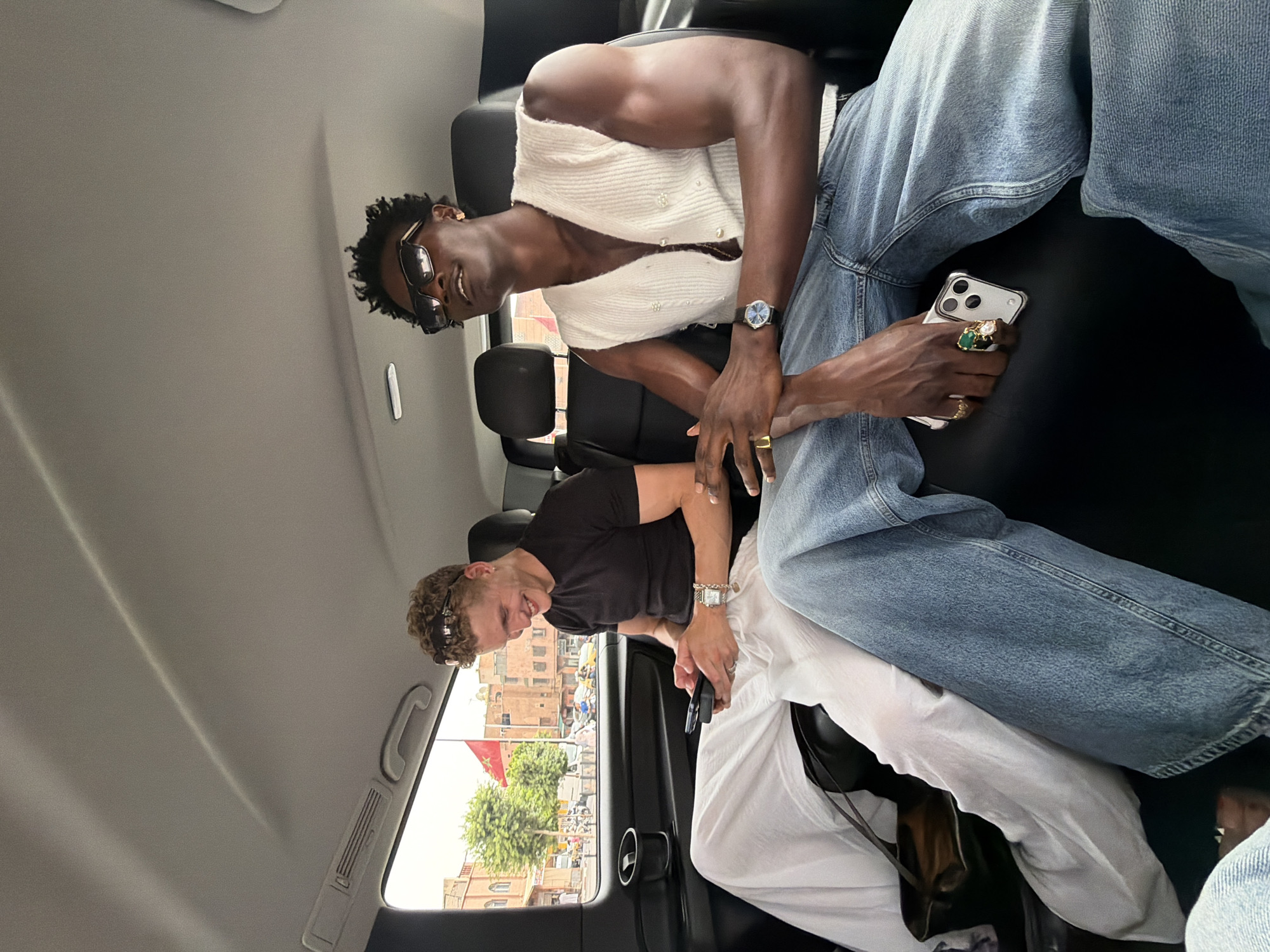 The media file captures a candid moment inside a vehicle, likely a car, with two individuals. The subject in the foreground is a Black man with dark curly hair, wearing sunglasses, a white open-buttoned vest over what appears to be a white garment, and blue denim jeans. He is looking towards the camera with a slight smile. His left hand, adorned with rings, is resting on his phone, which he is holding. A wristwatch is visible on his right wrist.
The second individual, partially visible in the background, is a lighter-skinned man with curly hair and sunglasses. He is wearing a black t-shirt and what appears to be white pants or a white garment. He is smiling and looking towards the camera. He also has a wristwatch on his right wrist.
The scene appears to be set during the daytime, with natural light filtering into the vehicle, suggesting fair weather. Through the car window, a bustling street scene is visible, with buildings that have a distinct architectural style reminiscent of Marrakesh, Morocco. Orange-hued buildings with arched windows are prominent, along with trees and a Moroccan flag. The overall atmosphere is relaxed and friendly, with the subjects appearing to be in good spirits. The text "MAROC" is visible on a flag.