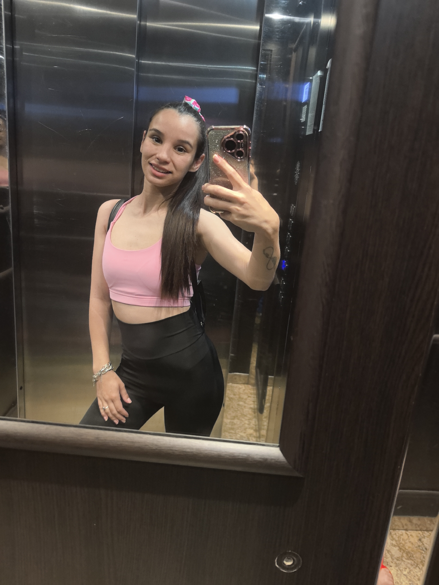 A young woman is captured in a mirror selfie taken inside an elevator. She stands facing the camera, holding a smartphone with a sparkly case in her right hand. She has long, dark hair tied up in a high ponytail with a pink and white scrunchie, and she is smiling faintly, revealing light-colored braces on her upper teeth.
Her attire consists of a pink sports bra and black high-waisted leggings. A black backpack is visible on her back. Accessories include a chunky silver-toned bracelet on her left wrist and a ring on her left ring finger. A small, abstract tattoo is visible on her left forearm.
The elevator interior features large, reflective metal panels on the back wall, reflecting some light and indistinct shapes. Dark wooden paneling or trim frames the mirror and forms the side wall on the right. An elevator control panel with illuminated blue buttons is faintly visible in the reflection to the right of the woman. The floor, visible in the reflection at the bottom, appears to have a light, patterned finish. The overall impression is a casual pose in a modern elevator setting.