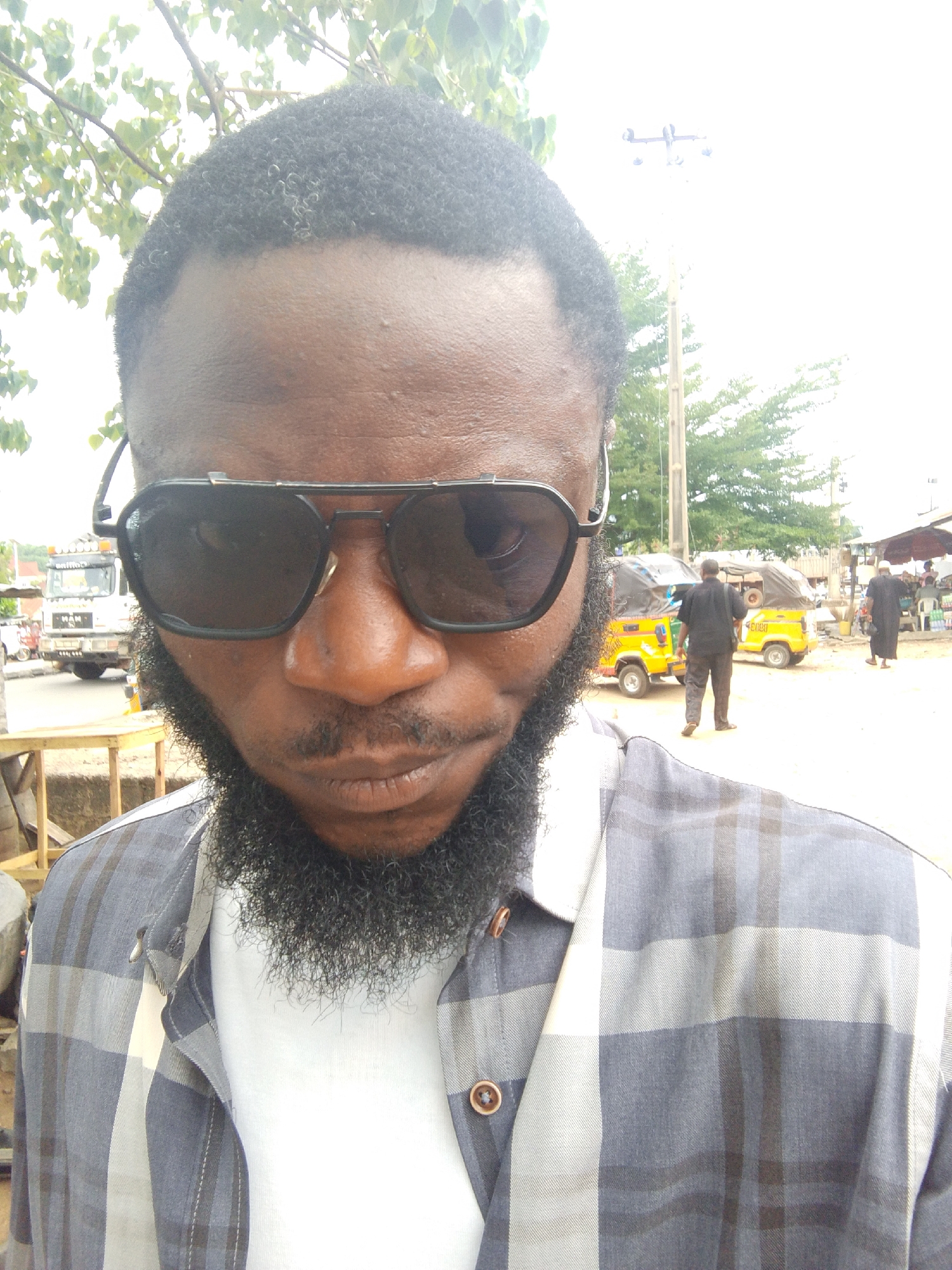 The image presents a close-up, slightly high-angle selfie of a dark-skinned man in an outdoor setting, likely Lokoja, Nigeria. The man, the primary subject, has a full black beard and mustache, along with short, dark curly hair. He is wearing large, dark aviator-style sunglasses that obscure his eyes, giving him a neutral or slightly serious expression. His attire consists of a white t-shirt beneath an unbuttoned grey and white plaid long-sleeved shirt.
The background reveals a bustling street or roadside scene under bright daylight. To the left, a white truck, identifiable by the "MAN" brand visible on its grille, is partially seen on a paved road, next to a wooden table or stand. To the right, an unpaved area features at least two yellow motorized tricycles, commonly known as "Keke Napep," with black canopies. Faint text, including "ENUGU," is discernible on one of the tricycles. Several people populate the background: one individual in dark clothing walks away from the camera, and two others, possibly in traditional robes, stand further back near what appears to be a roadside stall or shop. Green foliage from trees is visible in the upper corners and the distance, and a utility pole with wires stands on the right. The sky is very bright and appears somewhat washed out, indicating strong sunlight and clear weather during the day.