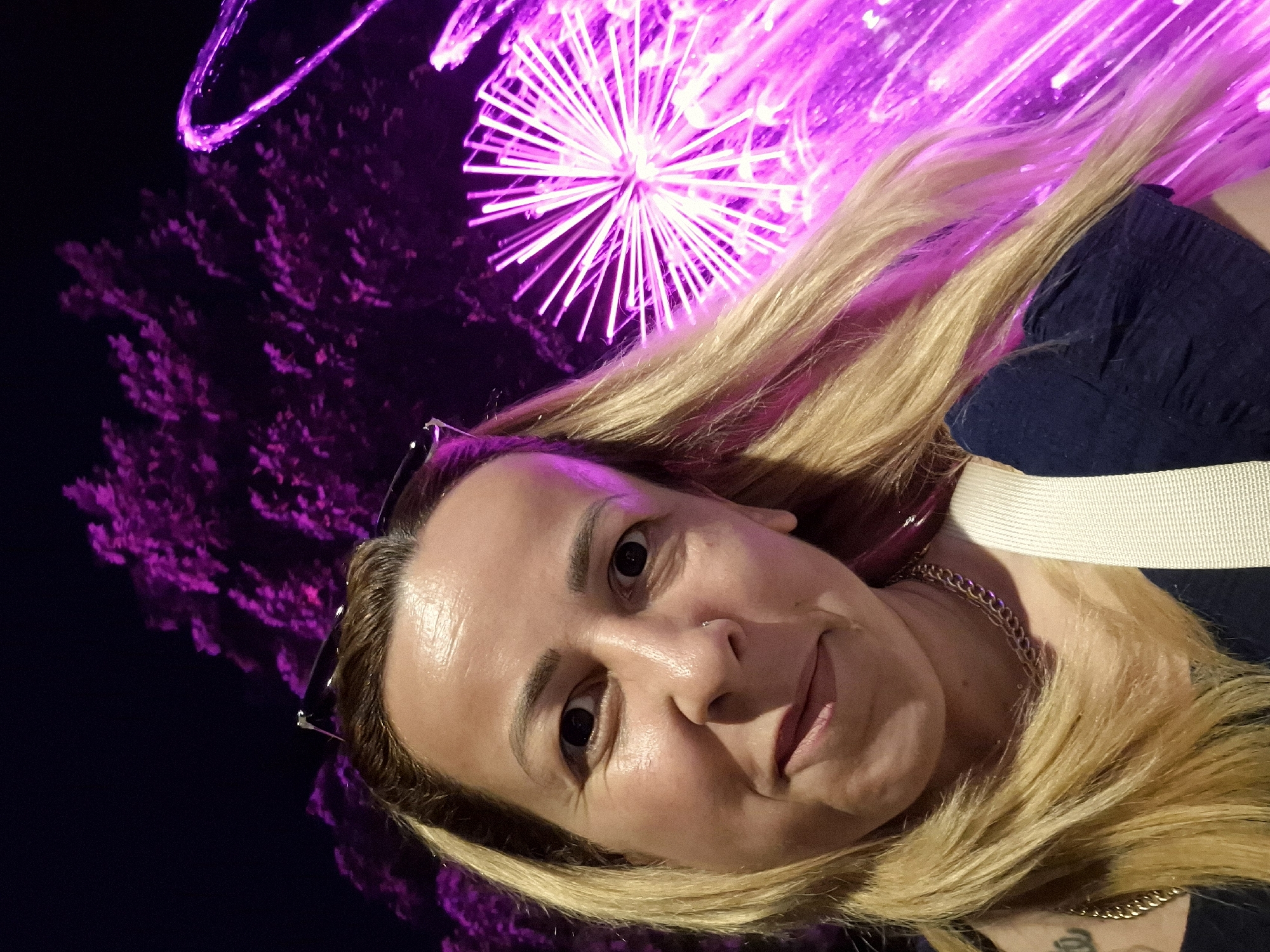 A woman with blonde hair smiles at the camera in a selfie. She is wearing a dark blue top and a gold chain necklace. Behind her, a fountain sprays pink water. The background is dark and blurry, with some pink lights visible. The location is not specified.