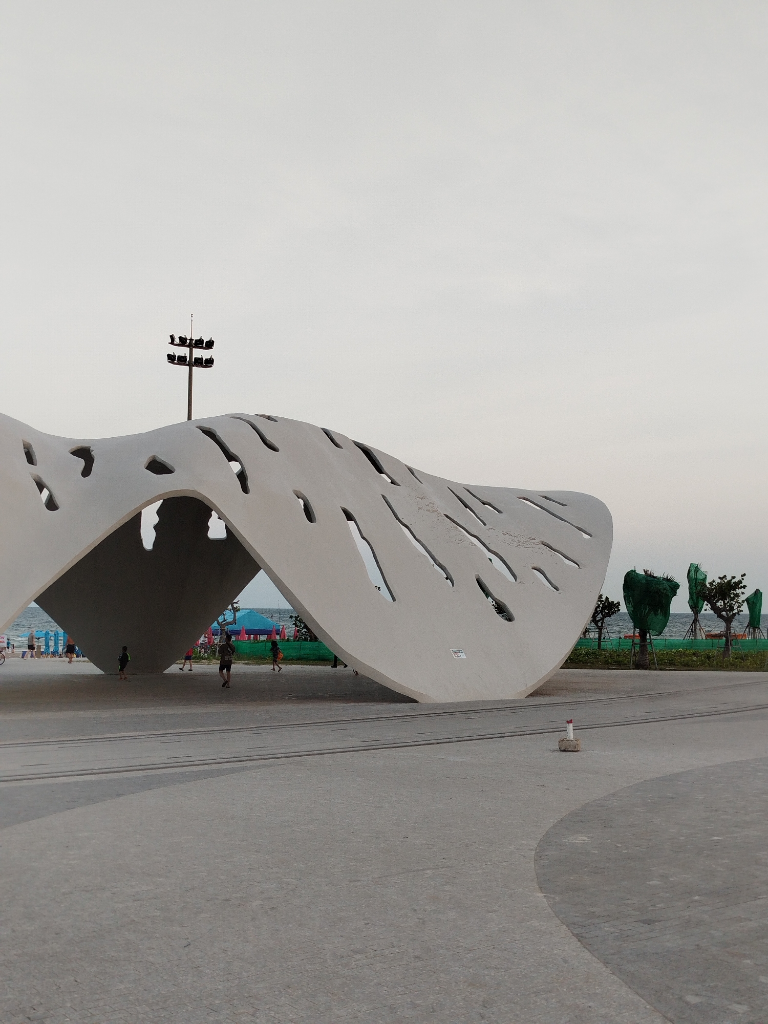 The photo depicts a large, modern art structure in a public space. The structure is white, with a curved, wave-like shape and several irregularly shaped openings. The structure appears to provide shade, as people are seen walking under it. In the background, there is a beach with people, trees, and a light pole. The ground is paved with grey stones. A small, concrete cylinder with a red and white top sits in the foreground. This location is likely somewhere in Ho Chi Minh City, Vietnam.