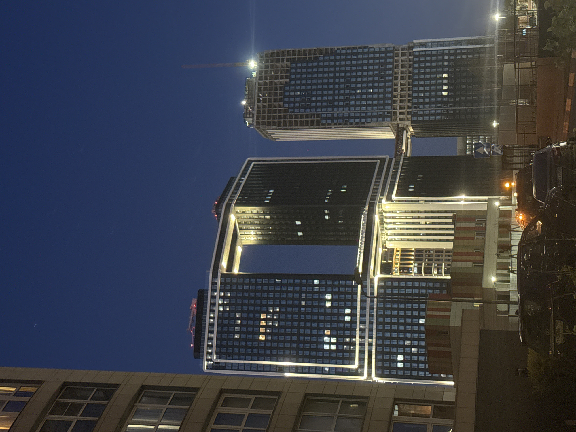 The image, captured at night or late dusk, showcases a modern urban architectural scene under a clear, dark blue sky.
The central focus is a distinctive, large U-shaped building complex in the mid-ground. It comprises two tall towers connected by a skybridge-like structure, creating a prominent rectangular opening in the middle. The building features dark facades with numerous illuminated windows, some forming visible patterns. White strip lighting accentuates the edges and structural lines of this modern complex.
Behind and slightly to the left of the U-shaped building stands another tall, rectangular skyscraper. This building appears to be under construction, with visible framework or unfinished sections on its upper floors. A tall construction crane, lit by work lights, extends from its summit.
To the right, additional illuminated building facades are visible, along with what appears to be a street or pathway. A dark vehicle, possibly a truck, is parked on the far right, with some faint, illegible text (potentially a license plate) visible on its rear. A blue and white road sign is also faintly discernible. In the foreground, at the bottom of the frame, sections of lower-rise buildings with windows are seen.
The scene conveys a sense of ongoing development and modernity in an urban environment, with the artificial lights of the buildings standing out against the dark sky. No people are visible.