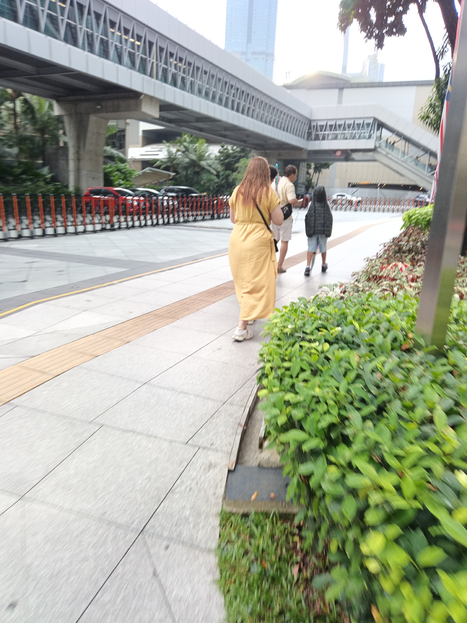 A group of people walk along a paved sidewalk adjacent to a lush green shrubbery. To the left, a series of red and orange bollards separates the sidewalk from a road where several cars are parked. An elevated walkway or bridge with a geometric design spans across the scene overhead. The environment appears to be an urban setting in Kuala Lumpur, Malaysia, possibly near a transportation hub given the elevated structure. The lighting suggests it is daytime, with overcast or diffused sunlight.