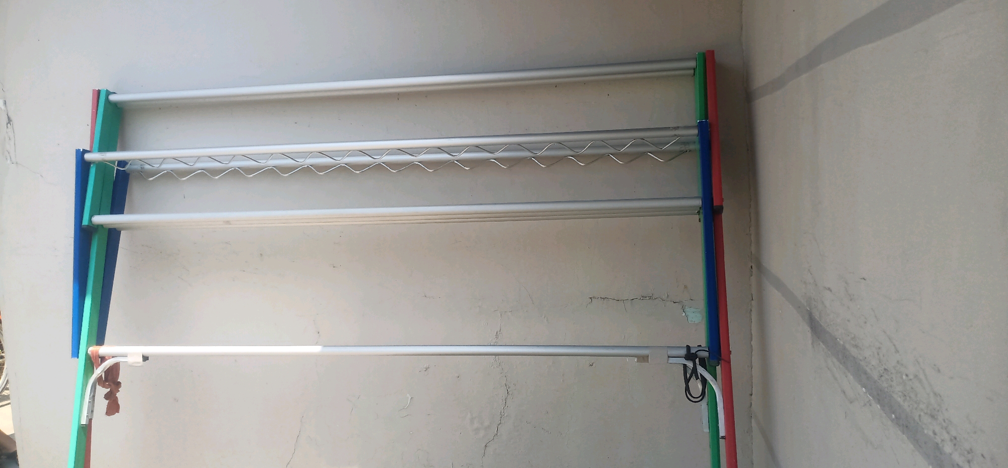 In Bajong, Indonesia, a multi-tiered drying rack stands against a textured concrete wall. The rack is constructed of silver-colored metal tubes and features colored vertical supports in red, blue, and green. The top two shelves are designed for hanging clothes, with the second shelf having a wavy, zig-zag pattern to potentially increase surface area for drying. The lowest shelf is a solid bar. A brown cloth is draped over the bottom shelf on the left side of the rack. The wall behind the rack shows signs of wear and tear, with visible cracks and some discoloration. The lighting suggests an outdoor setting, possibly a patio or courtyard, with shadows cast on the wall.