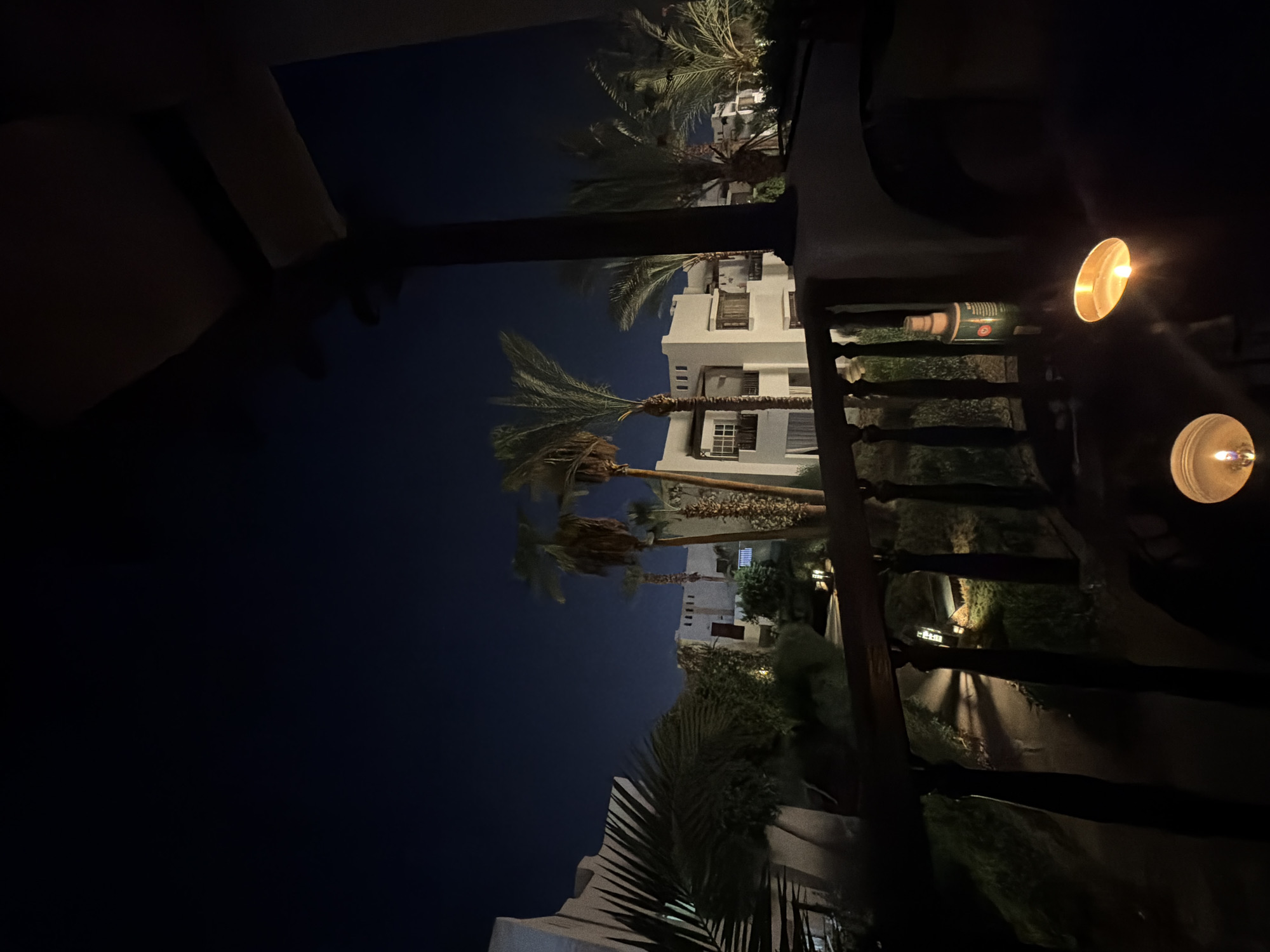 The image shows a nighttime view from a balcony overlooking a resort in Sharm El Sheikh, Egypt. White stucco buildings with multiple windows and balconies are visible, surrounded by lush greenery and palm trees. The sky is a deep, dark blue. On the balcony in the foreground, two lit candles are present, casting a warm glow on the scene. A wooden railing separates the balcony from the garden area below, where pathways are illuminated by soft lights. A green bottle, possibly a cleaning or repellent spray, stands on the railing. The overall atmosphere is serene and tranquil, suggesting a peaceful evening at a tropical resort.