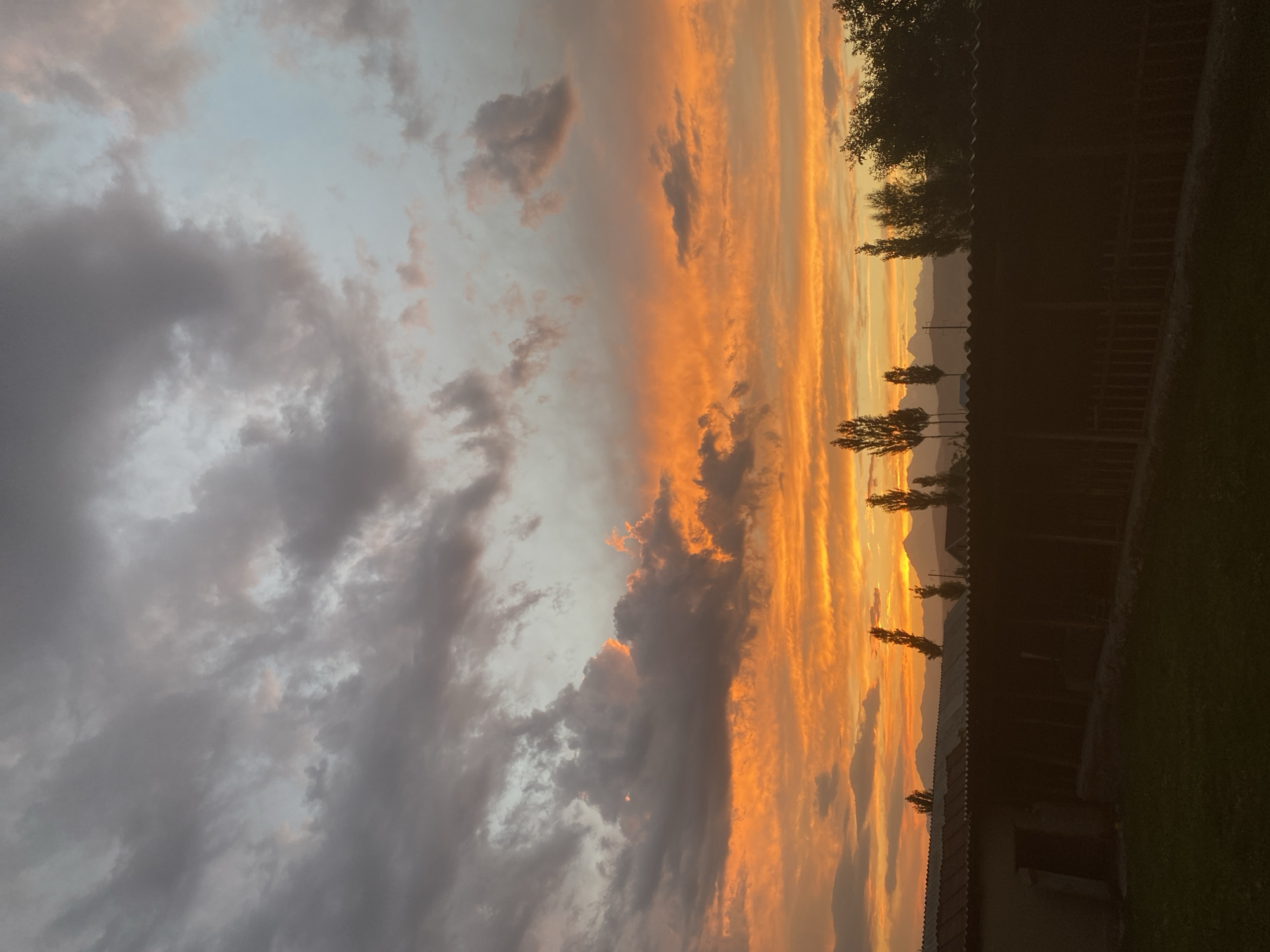This is a vertically oriented, eye-level, full shot of a sunset. The sky is filled with dramatic clouds, transitioning from dark gray on the left to vibrant oranges and yellows on the right. The sun is low on the horizon, casting a warm glow through the clouds. In the background, silhouetted mountains are visible. In the foreground, the top of a dark, rustic building with a corrugated roof runs horizontally across the frame, with a wooden fence along its edge. A few trees with dark foliage are also silhouetted against the colorful sky. The overall mood is serene and awe-inspiring, evoking the beauty of nature at the end of the day. The time of day is clearly sunset, and the weather appears to be partly cloudy. There is no visible text in the image.