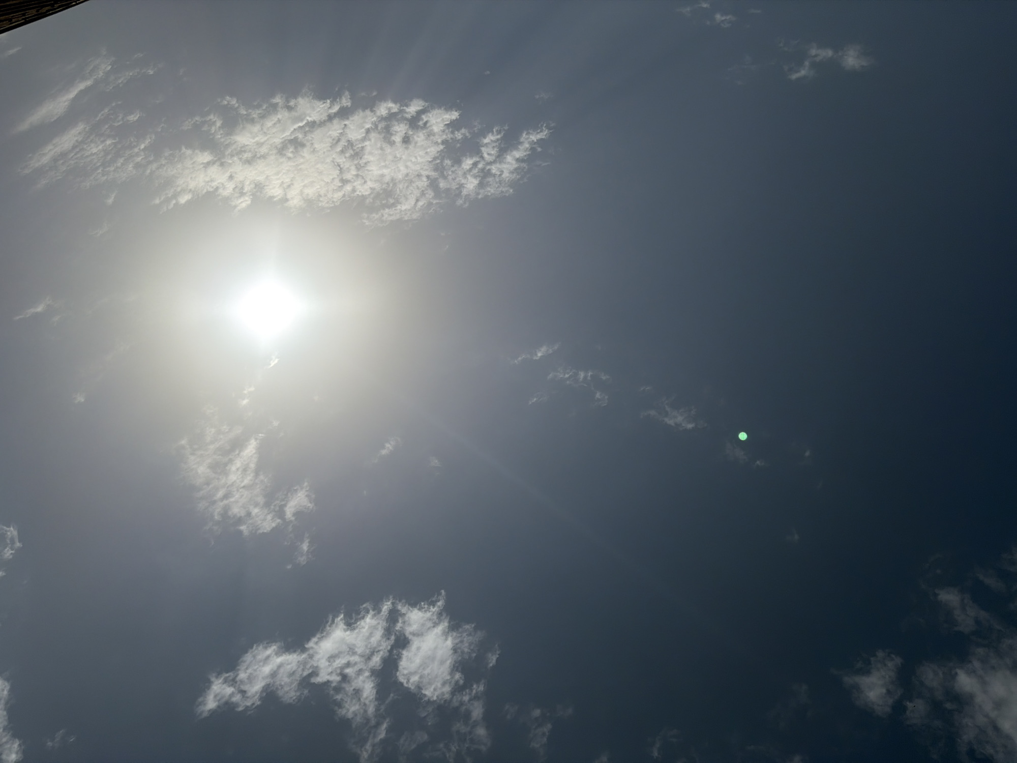 The image depicts a bright sun, appearing as a square of light, positioned in the upper center of a dusky blue sky. Wispy white clouds are scattered across the sky, with some appearing more dense than others. A small, glowing green dot is visible in the lower center of the frame, and a hint of a dark structure, possibly a building or tree, is present at the very top edge of the image. The lighting suggests it is daytime, possibly late afternoon or early morning, given the intensity and position of the sun. The scene appears to be an outdoor view, looking directly upwards. There are no people or animals visible in the image. The scene is devoid of any specific landmarks or elements that would definitively place it in Jos, Nigeria, beyond the provided context.
