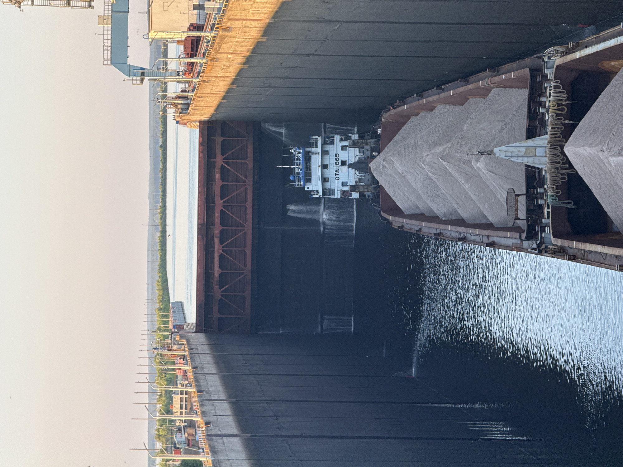 This media file captures a scene aboard a large cargo ship filled with what appears to be a load of gravel or sand. The vessel is positioned on a body of water, likely a river or canal, with its vast, dark deck stretching out of frame. The material is piled high in distinct, sloping mounds within the ship's cargo holds.
A smaller vessel, a tugboat or service craft, is positioned between the cargo ship and a section of the port infrastructure visible on the left. This smaller boat is white with blue accents and has the designation "OTA 865" clearly visible on its side. The water surrounding the vessels is dark and slightly choppy, reflecting the overhead structures and the sky.
The time of day appears to be late afternoon or early evening, suggested by the warm, soft light casting long shadows across the deck of the cargo ship. The sky is a hazy, pale orange, indicating a possible sunset or atmospheric conditions like dust or light fog. The port infrastructure on the left includes industrial structures with walkways, cranes, and supporting beams, hinting at an active shipping or industrial zone.
The overall impression is one of industrial activity and transportation on a large scale. The focus is on the immense size of the cargo ship and its payload, with the smaller tugboat providing a sense of scale and movement within the scene.