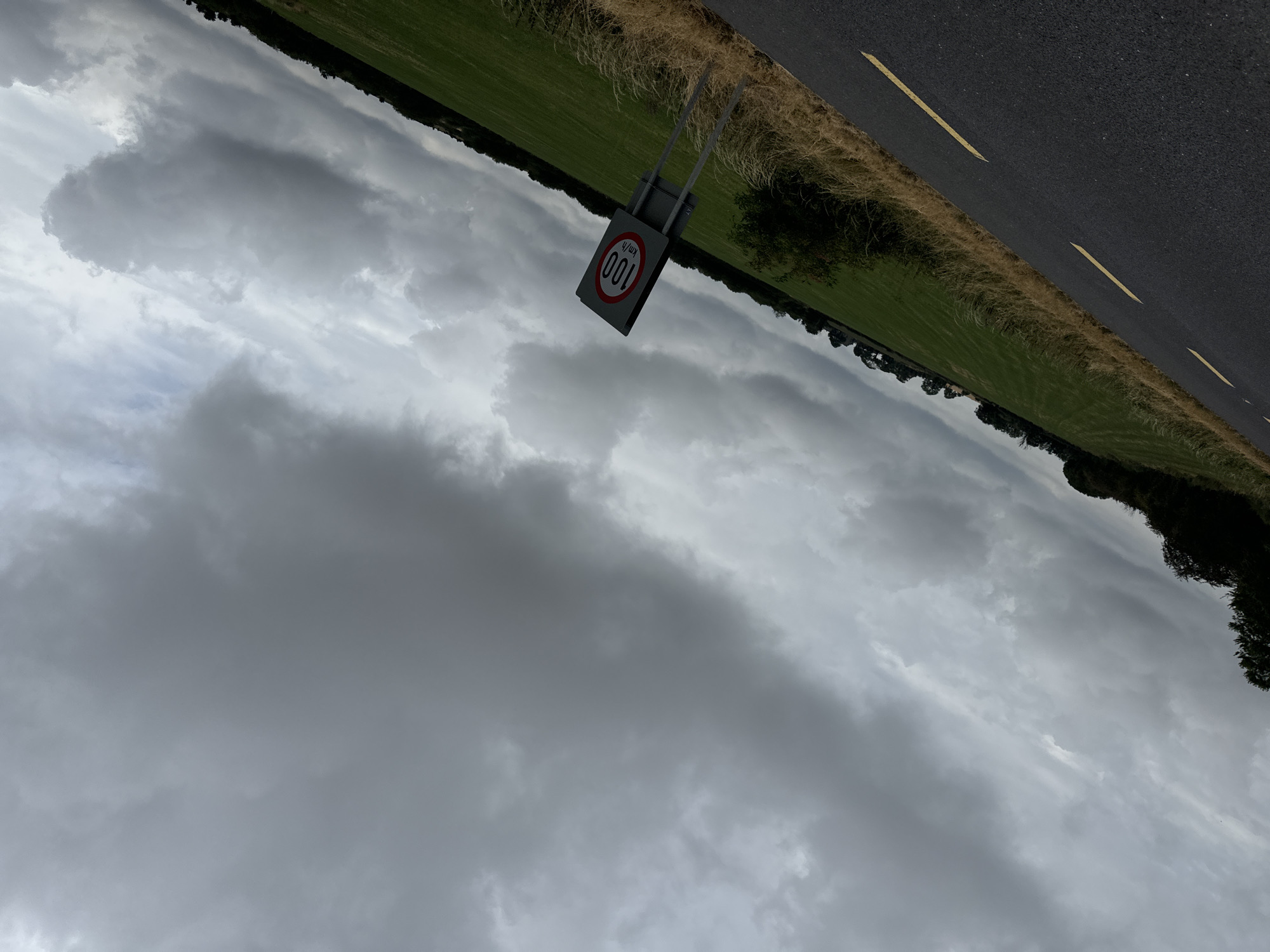 The image depicts a scene on a road, likely in Kilivory, Ireland, given the context. The road is paved with asphalt and has yellow dashed lines indicating lanes. The sky is heavily clouded with dark, dramatic storm clouds. To the right of the road, a speed limit sign is visible, showing "100 km/h" within a red circle, mounted on two metal poles. Beyond the road and the sign, a vast expanse of green farmland stretches towards a line of trees in the distance. The land is a vibrant green, suggesting it is well-maintained pasture or fields. The overall atmosphere is overcast and somewhat ominous due to the thick cloud cover. There are no people or animals visible in the frame. The composition is at an angle, giving a sense of movement or perspective from a vehicle traveling along the road.