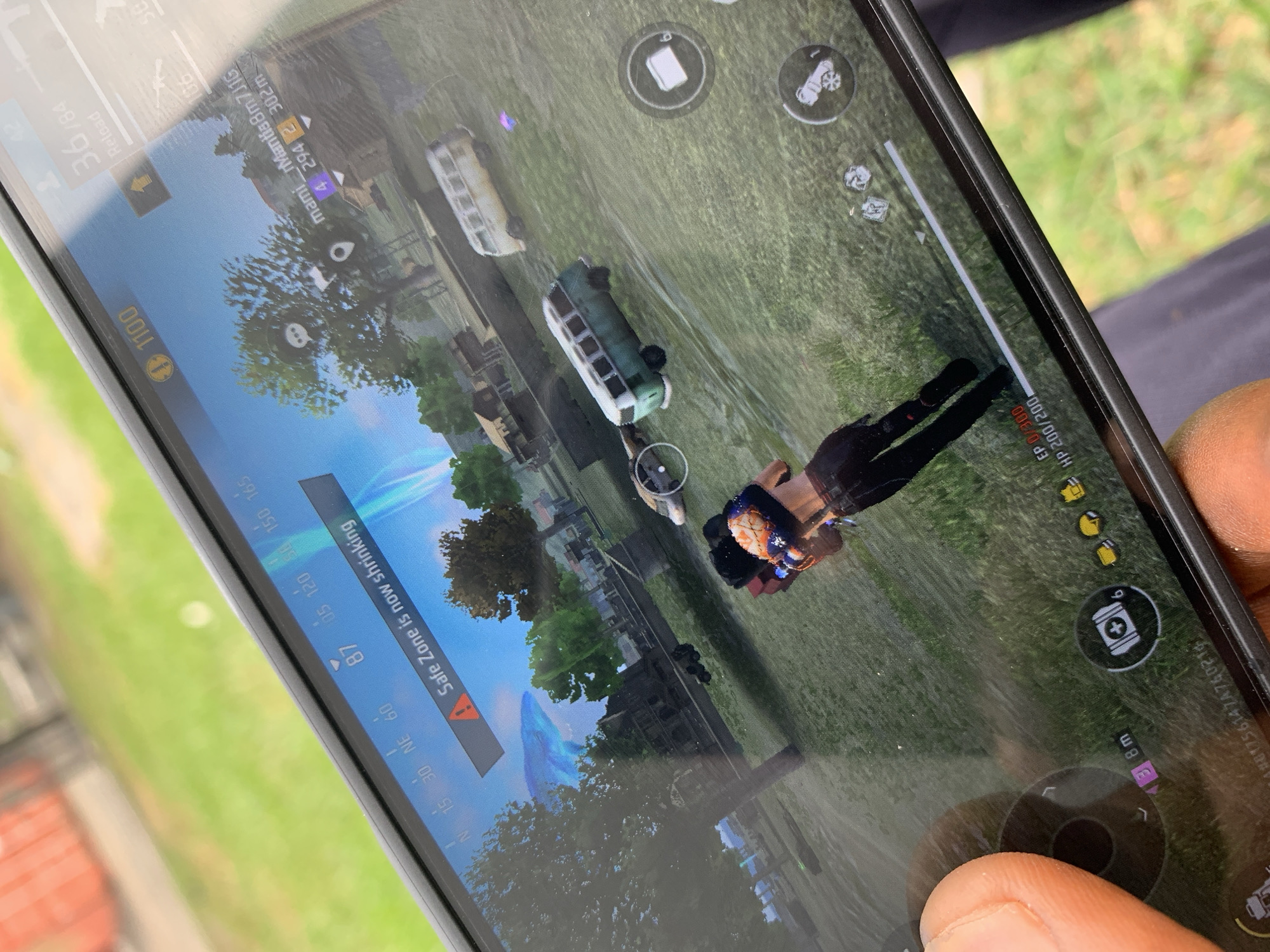 A close-up, angled shot captures a mobile phone screen displaying gameplay footage from what appears to be a battle royale-style video game. The game environment depicts a green, grassy area with scattered vehicles, including buses. A player character is visible, positioned centrally on the screen. Game interface elements such as player statistics (HP/EP), currency ("1100"), compass direction (NE), and distance to the safe zone are displayed. A person's fingers are partially visible, suggesting they are operating the phone. The surrounding environment is out of focus, showing greenery suggesting an outdoor setting. The location is likely Sangotedo, Nigeria, based on the presence of the person, however, it can only be inferred through contextual clues from the image as there's no visible evidence of the city.