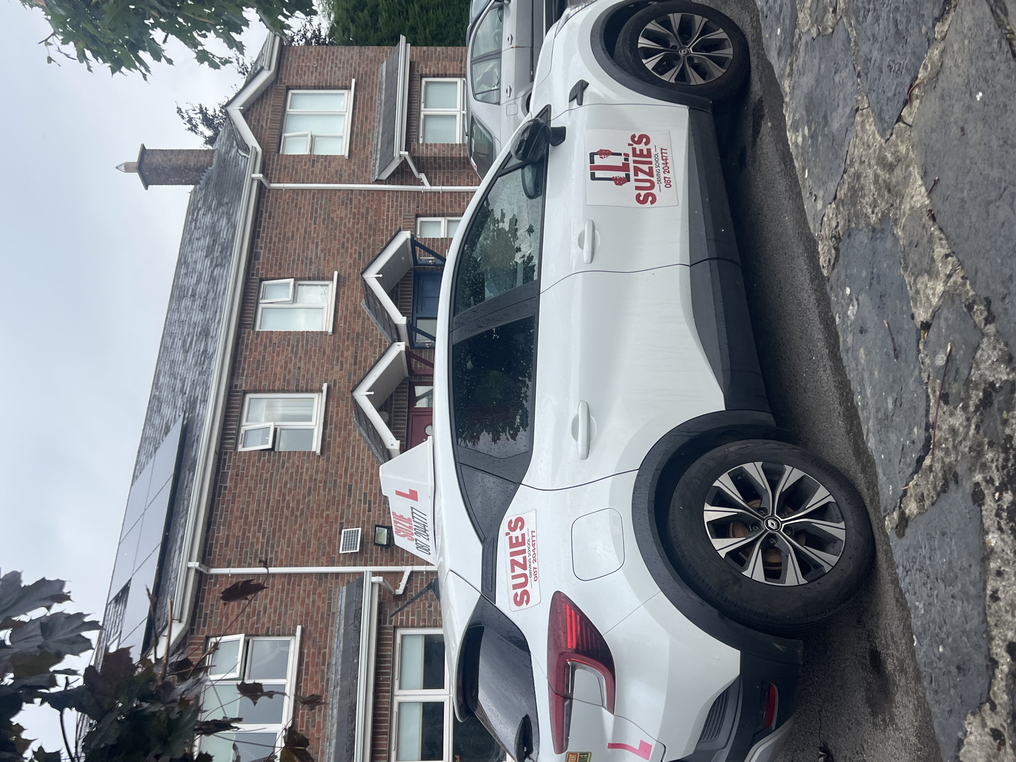 A white, four-door hatchback driving school car with "SUZIE'S DRIVING SCHOOL" decals is parked on a paved surface in front of a red brick two-story house. The car has a "L" plate on the roof and a pink "L" on the rear left fender. The house has multiple windows, a chimney, and solar panels on the roof. A silver sedan is parked in front of the house to the right of the driving school car. The image is taken from a low angle, with tree branches visible in the upper left corner. The ground appears wet, possibly from rain.