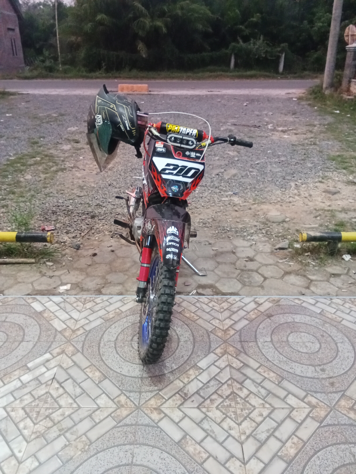 A black and green motocross helmet with a visor is hanging from the handlebars of a motocross bike. The bike is black, red, and white with the number 210 displayed prominently on the front. It has knobby tires suitable for off-road use and is parked on a tiled patio. In the background, there is a gravel-covered area, a road, and vegetation, suggesting a rural or semi-rural setting. The scene appears to be in Jirak, Indonesia, likely in front of a house or a roadside establishment given the tiled patio and the presence of a building on the left. The lighting indicates it may be late afternoon or early evening.