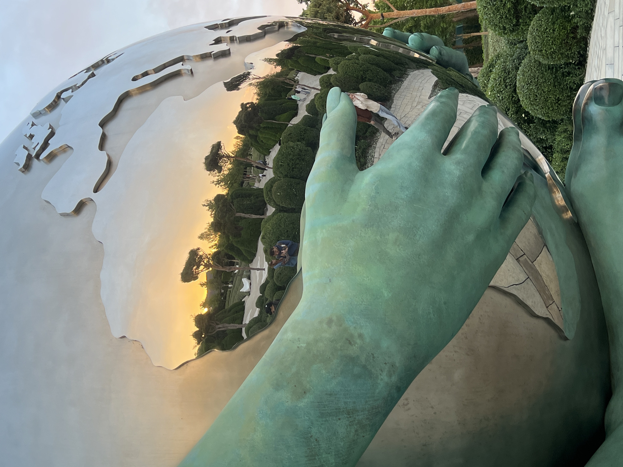 This image captures a monumental sculpture featuring a highly reflective, metallic globe embraced by large, patinated green hands and feet, likely made of bronze. The smooth, mirror-like surface of the globe prominently reflects its surroundings, creating a panoramic view.
In the reflection, a warm, golden glow suggestive of either sunrise or sunset illuminates the horizon and the lower portion of the globe. Lush green trees, bushes (some appearing manicured), and a paved pathway are clearly visible in the distorted reflection. Several people are also reflected on the globe's surface; at least two individuals in casual attire can be seen walking on the path, observing the scene. The sky above the reflected landscape appears bright and clear.
The sculpture's green hands and feet, showing signs of weathering or patina, are positioned as if cradling or resting on the polished globe. The overall setting appears to be an outdoor public space, likely a park or plaza, characterized by landscaping and pedestrian pathways, consistent with locations such as the "Planet of Children" sculpture in Krasnodar, Russia.
The photograph is taken from a close, low-angle perspective, emphasizing the scale of the sculpture. No discernible text is visible in the image. The scene conveys a sense of peaceful contemplation under the warm glow of the sun.