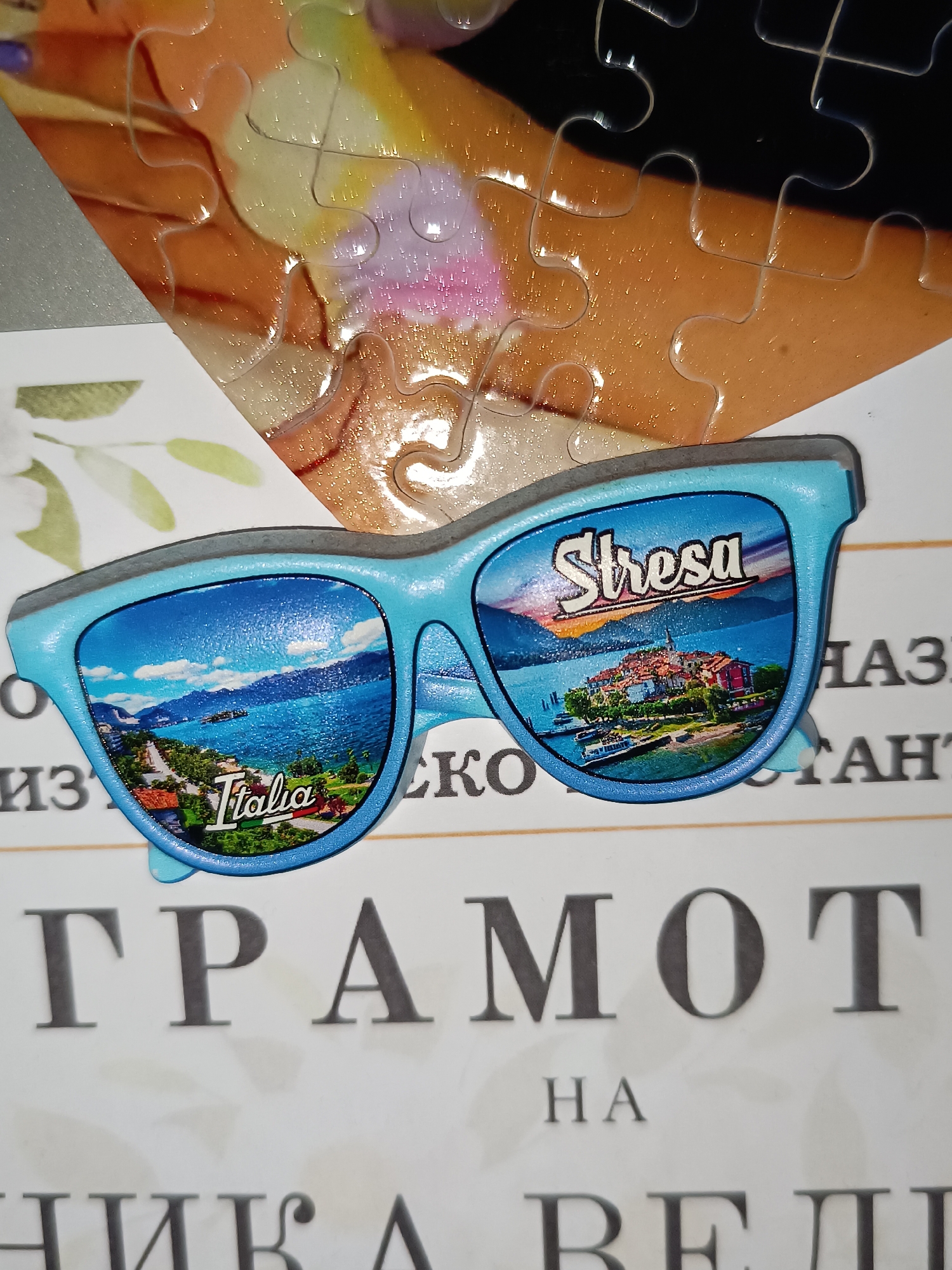 The image shows a pair of light blue sunglasses with images of Italy, specifically the town of Stresa, printed on the lenses. The sunglasses are placed on a document written in Bulgarian. The document appears to be a certificate or diploma, judging by the large lettering which includes the word "ГРАМОТ" (Gramota, which translates to Diploma/Certificate). A portion of a jigsaw puzzle is visible in the background. There is no indication of the location being the city of Pleven, Bulgaria.